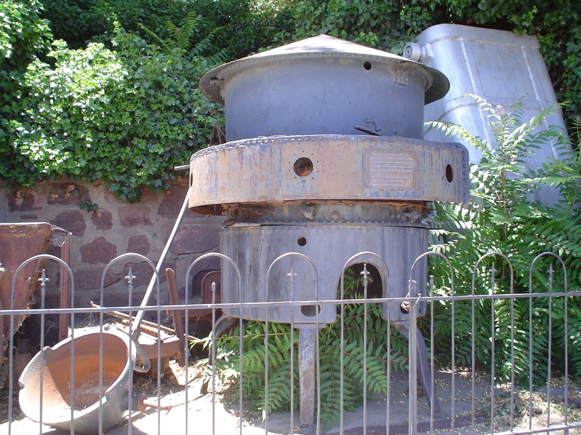 A large metal object is behind a fence with a few plants in the background