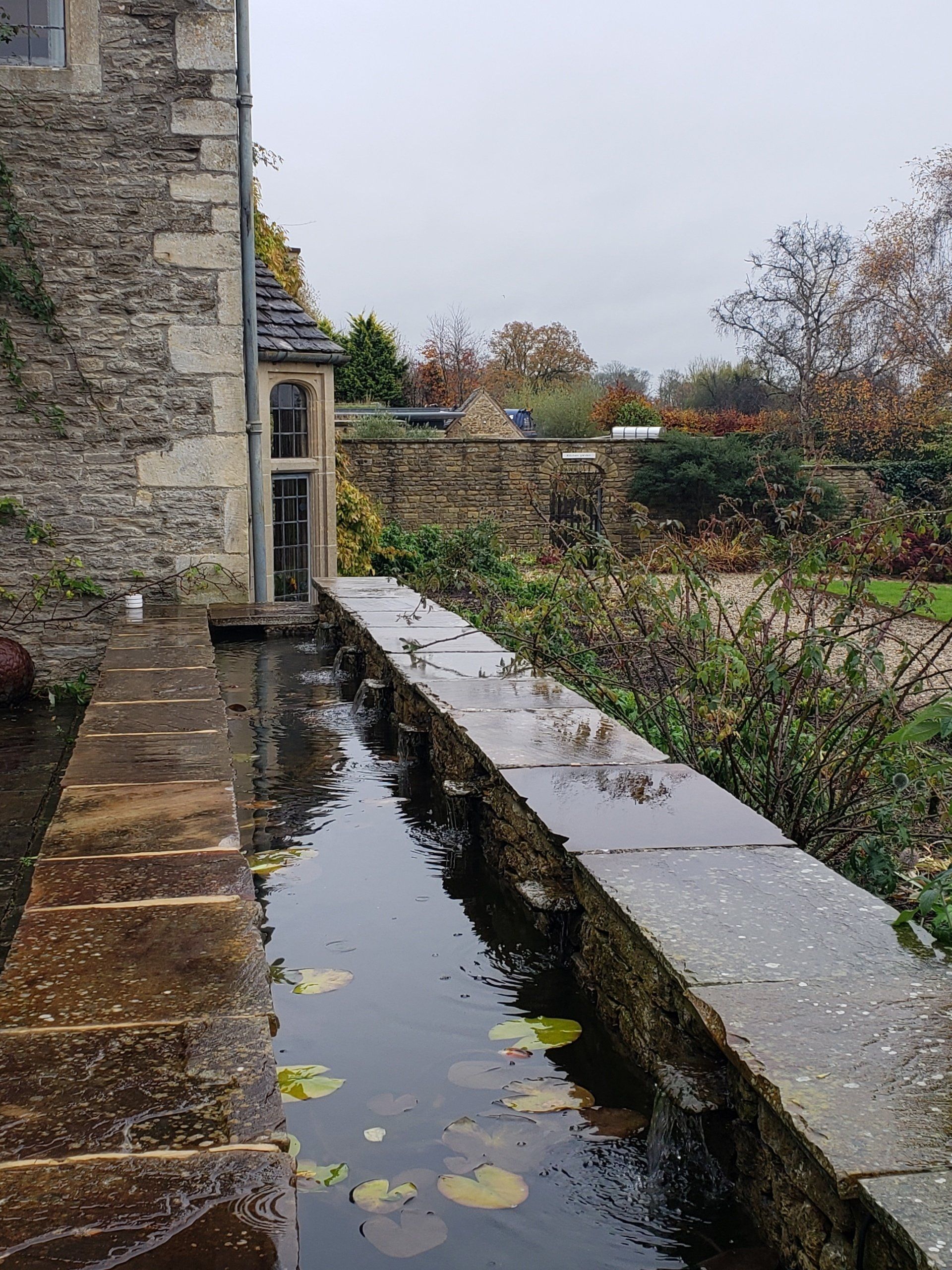 There is a pond in the middle of a garden next to a stone wall.