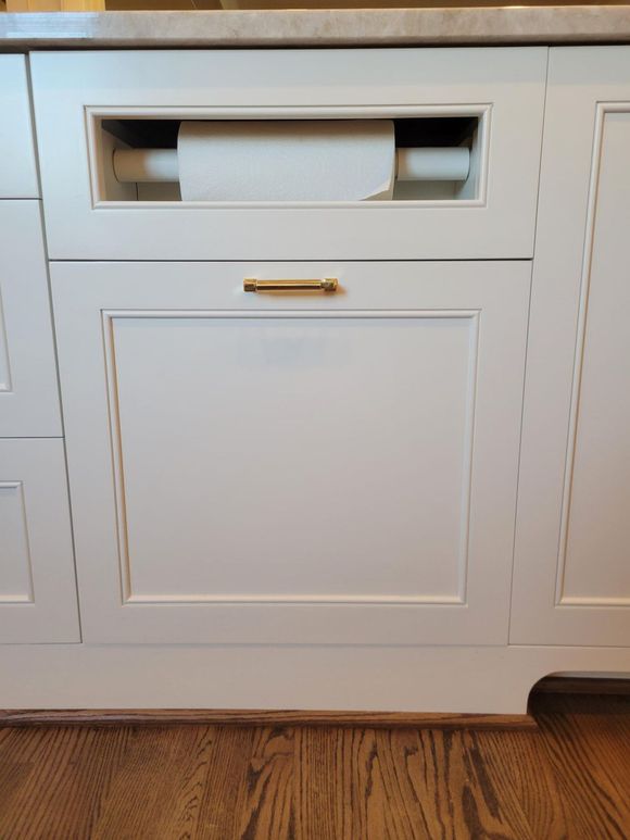 White cabinet with a built-in pull-out paper towel holder and brass handle on a wood floor.
