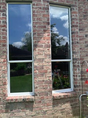 Two tall, narrow windows in a brick wall reflect the sky and a yard with greenery.