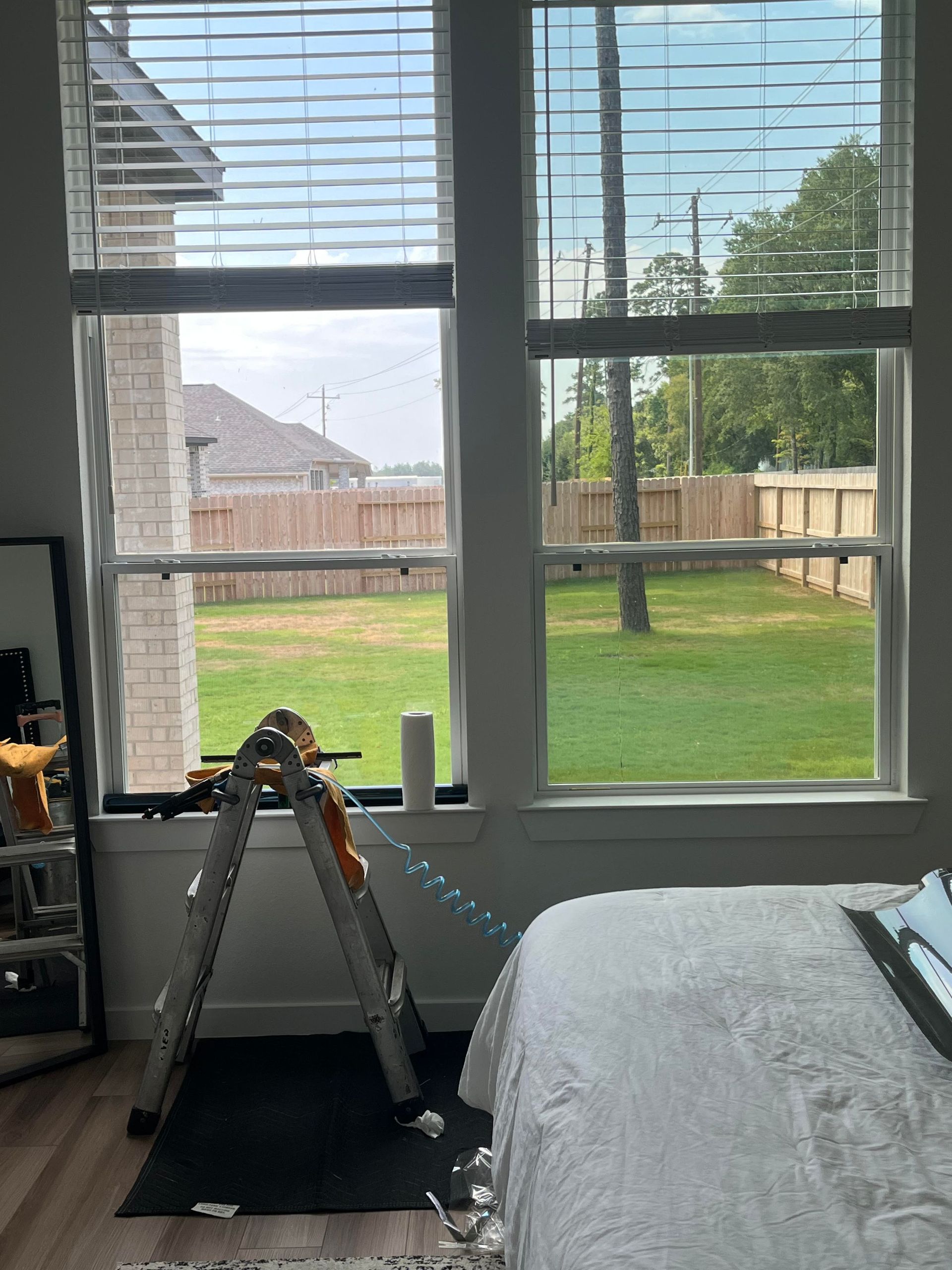 Interior view: tall windows with blinds overlooking a backyard, ladder propped inside near bed.