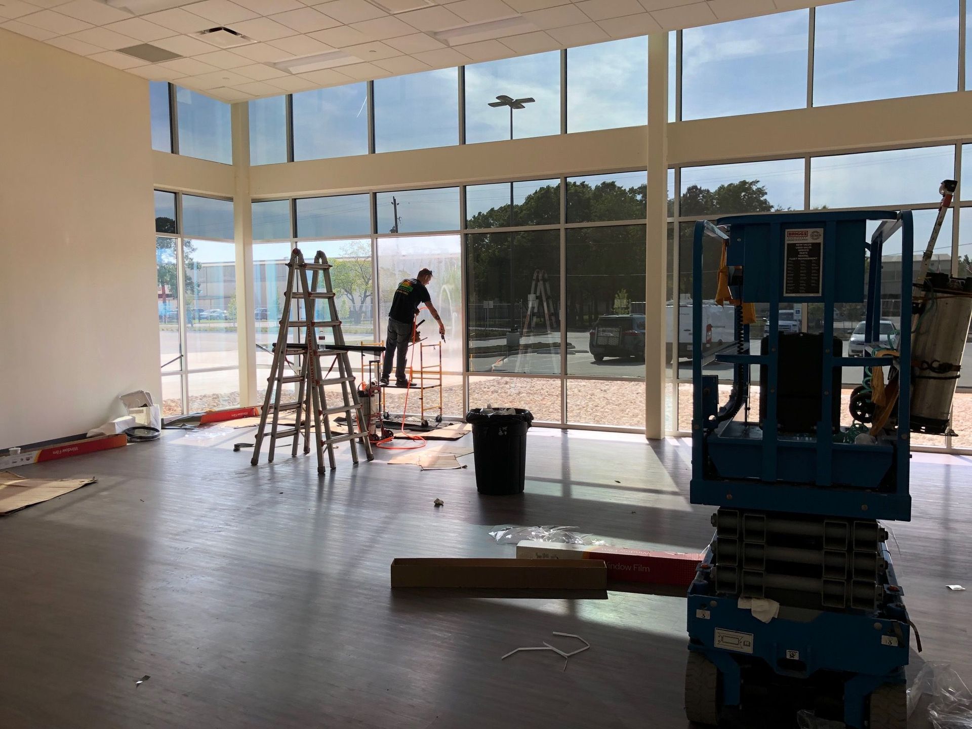 Interior view of a building under construction. A person works on large windows, using a lift and a ladder.