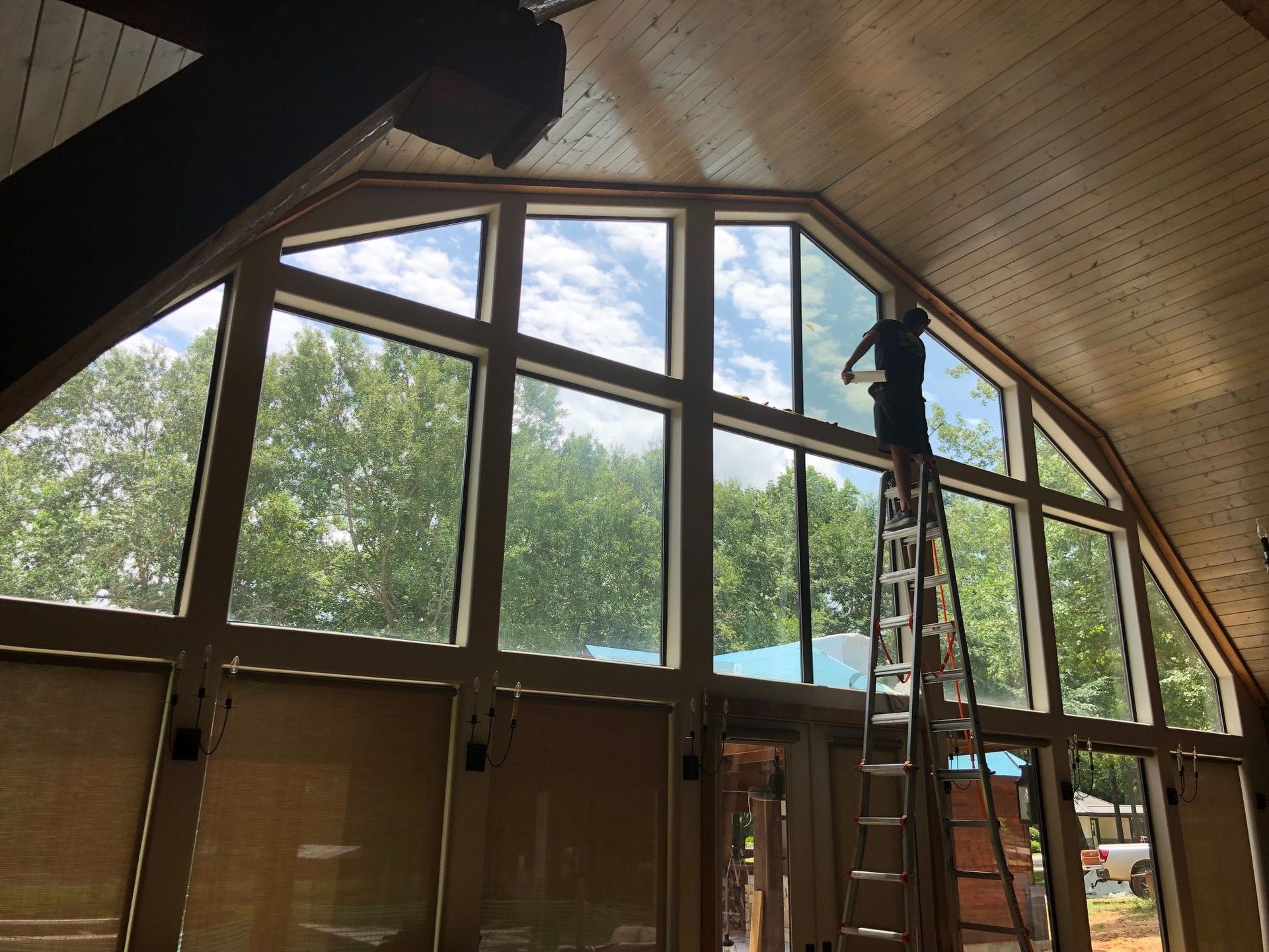 Person on a ladder cleaning a large window with trees and sky visible through it. Interior with wood ceiling.
