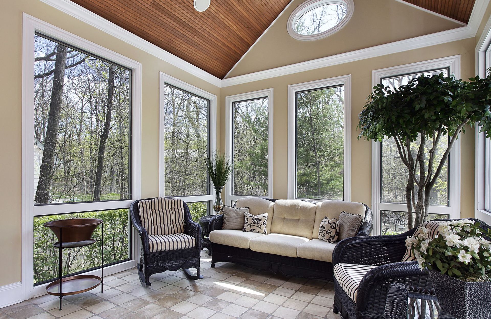 Sunroom with wicker furniture, large windows overlooking trees, beige walls, and wood ceiling.