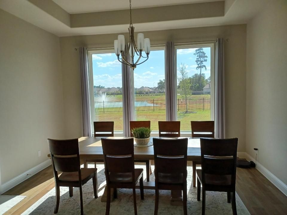 Dining room with large windows overlooking a pond. Table with eight chairs, a chandelier, and light gray curtains.