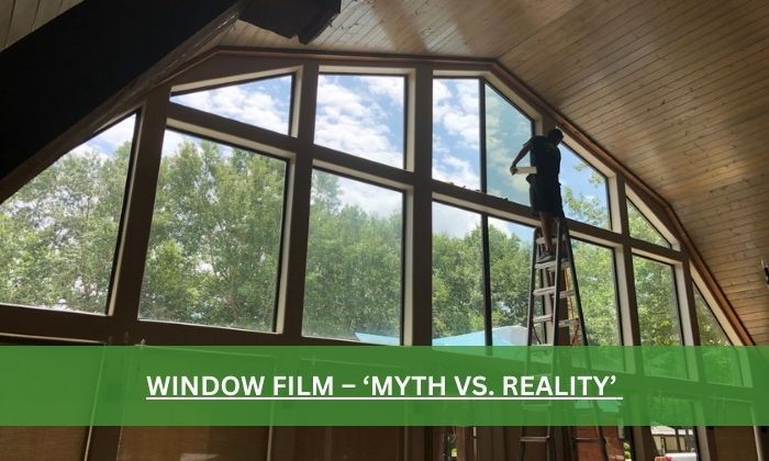 Person on a ladder installing window film inside a house with large windows and wooden beams.