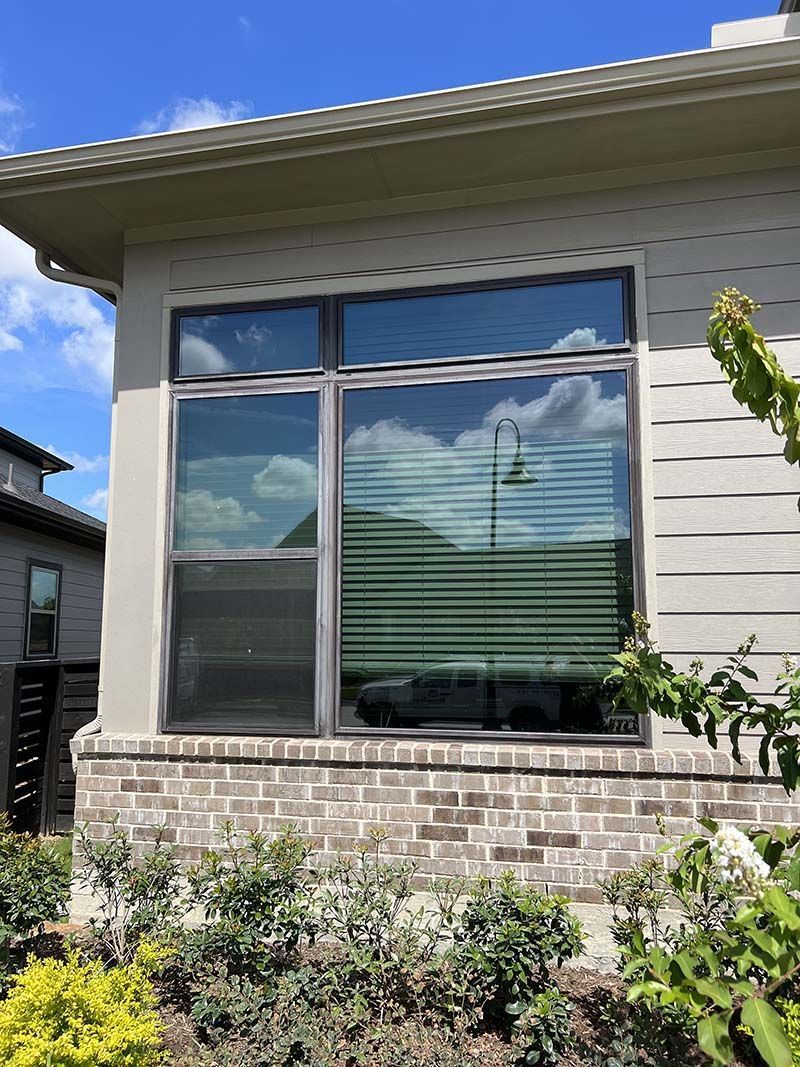 Window of a house with dark frames, reflecting clouds and blinds. Brick base with bushes and a sunny sky.