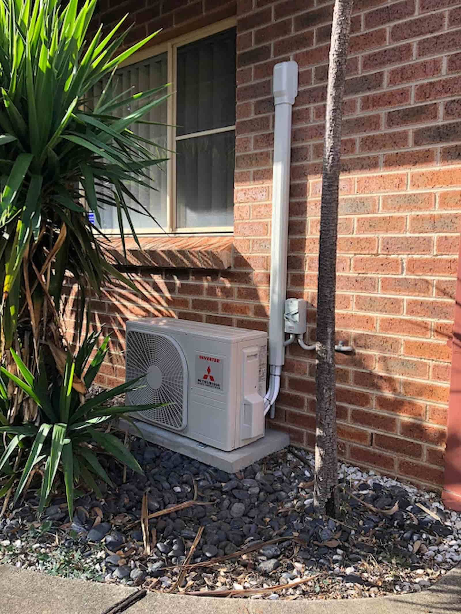 A White Air Conditioner is Sitting on the Side of a Brick Building Next to a Tree — Steve's Air Conditioning in Lakeside Dr, NSW