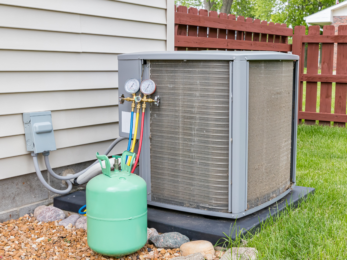 An air conditioning unit outdoors with gauges attached, and a refrigerant tank nearby.
