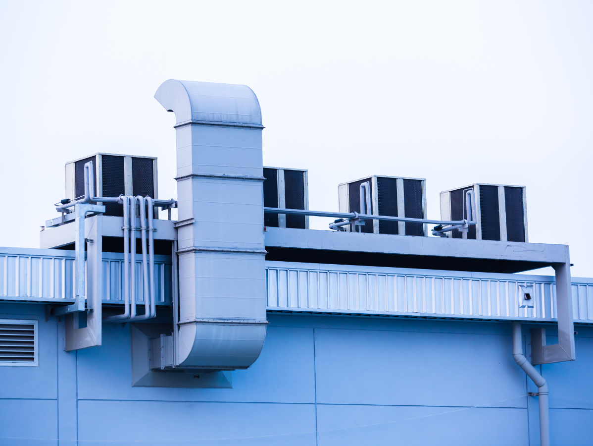 Air conditioning units and ductwork on a light blue building's roof against a pale sky.