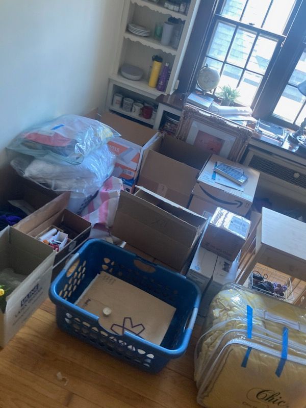 Boxes and laundry basket in a room, likely moving. Clutter with bags, containers, and a window.