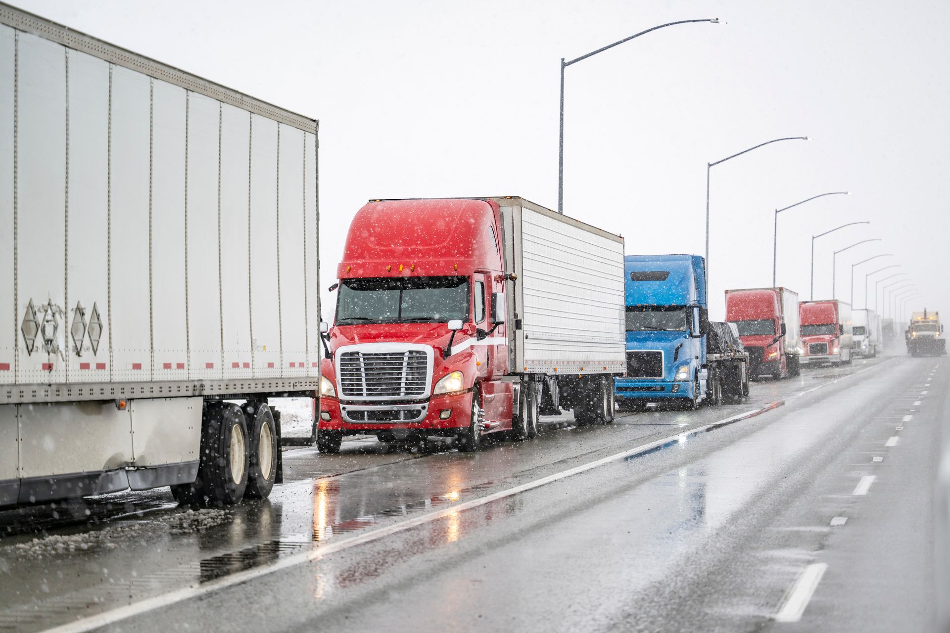 Semi-trucks in a snowy setting on a wet highway. Red, white, and blue trucks in a line.