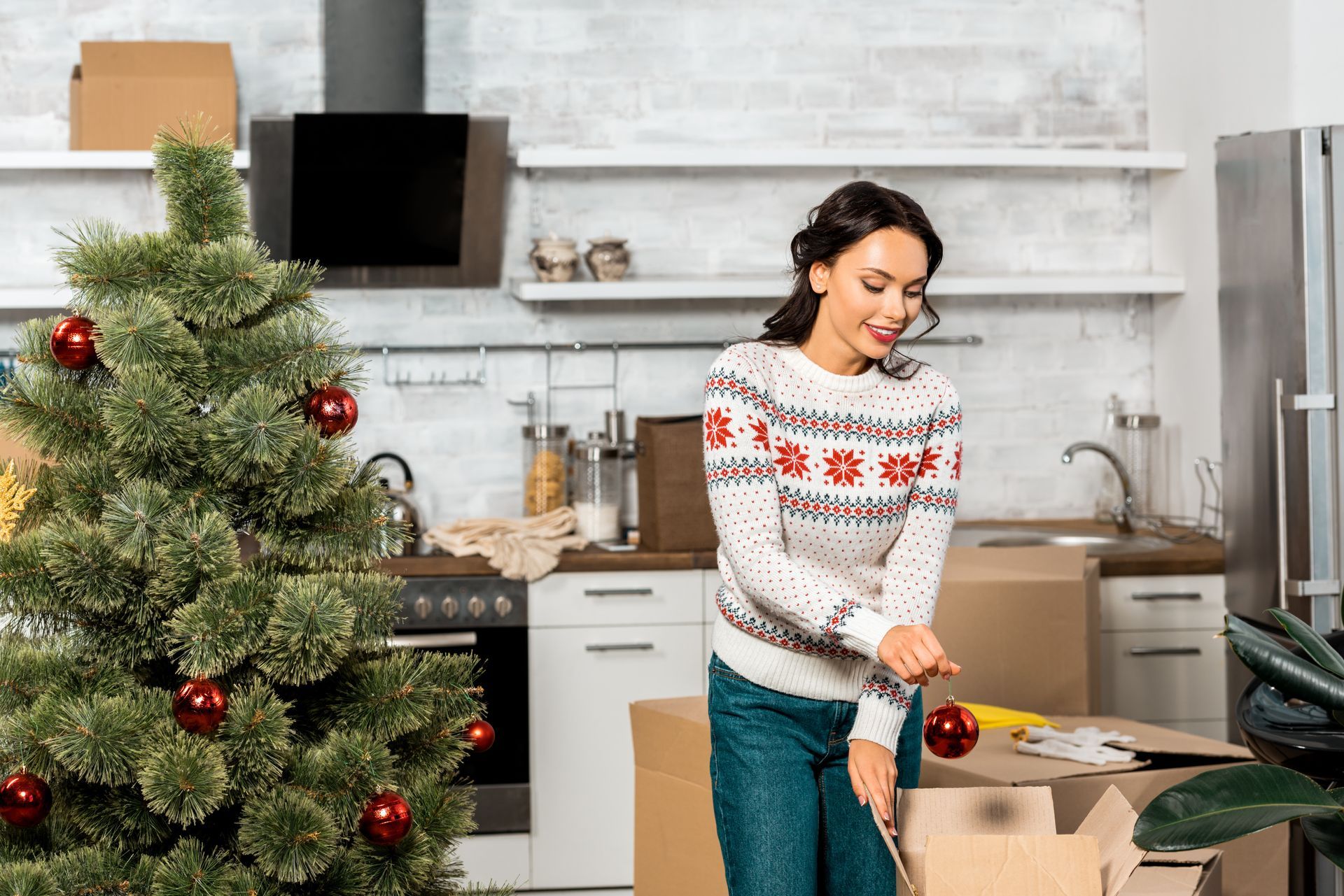 Woman decorating a Christmas tree in a kitchen with moving boxes.