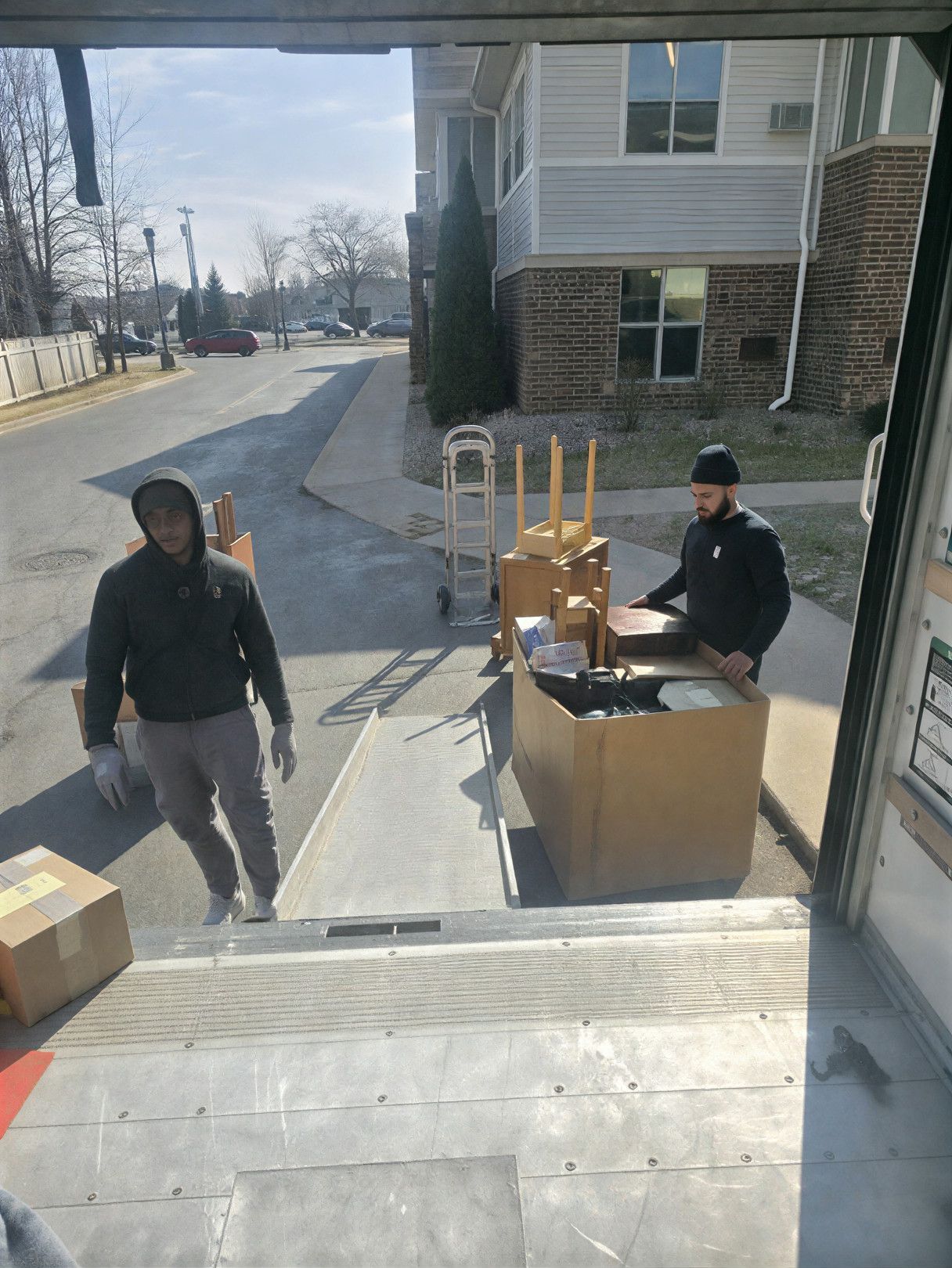 Two movers loading boxes from a truck ramp. One walks away, the other stands. Sunny day, apartment building in background.