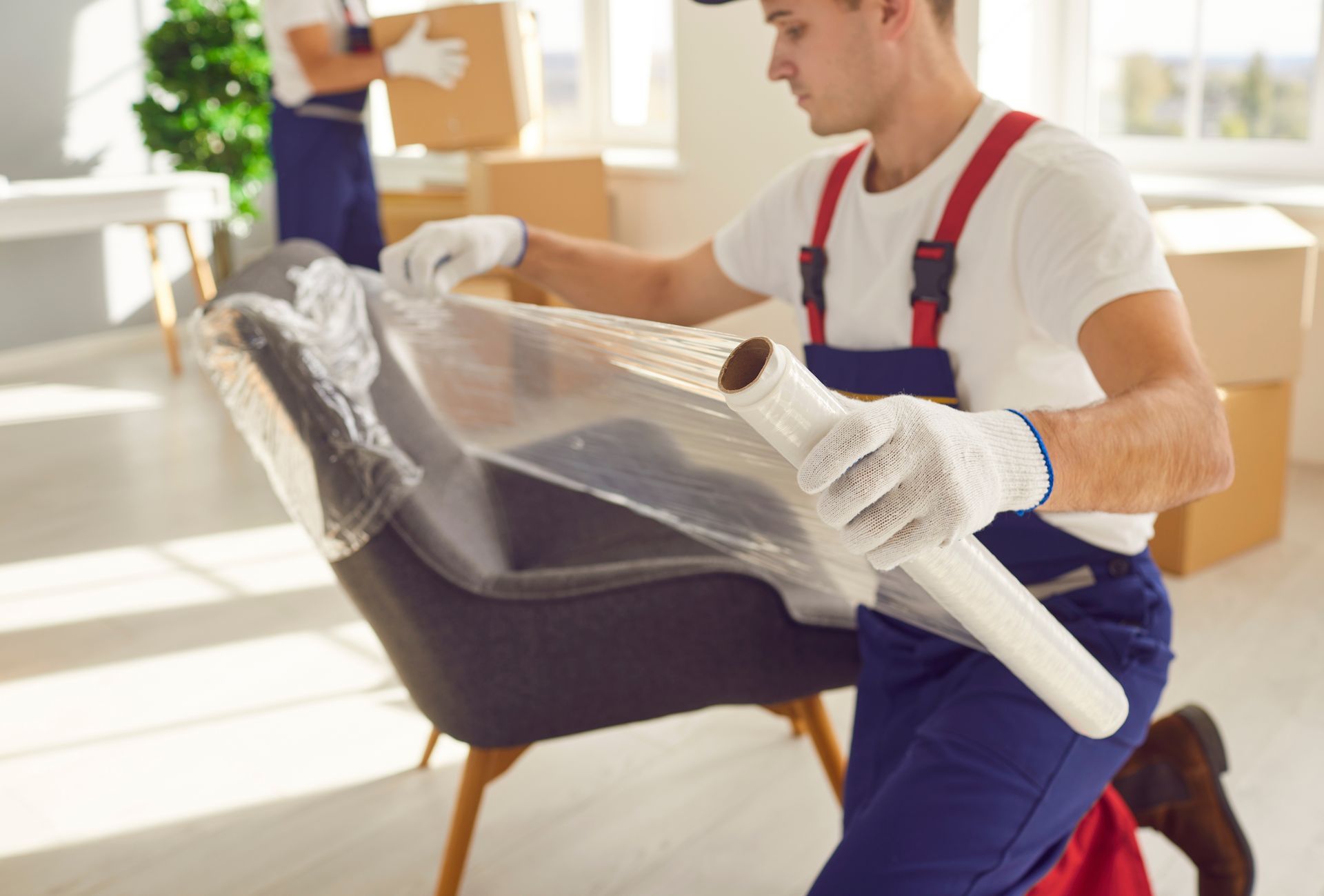 Movers wrapping a chair in plastic in a room with boxes.