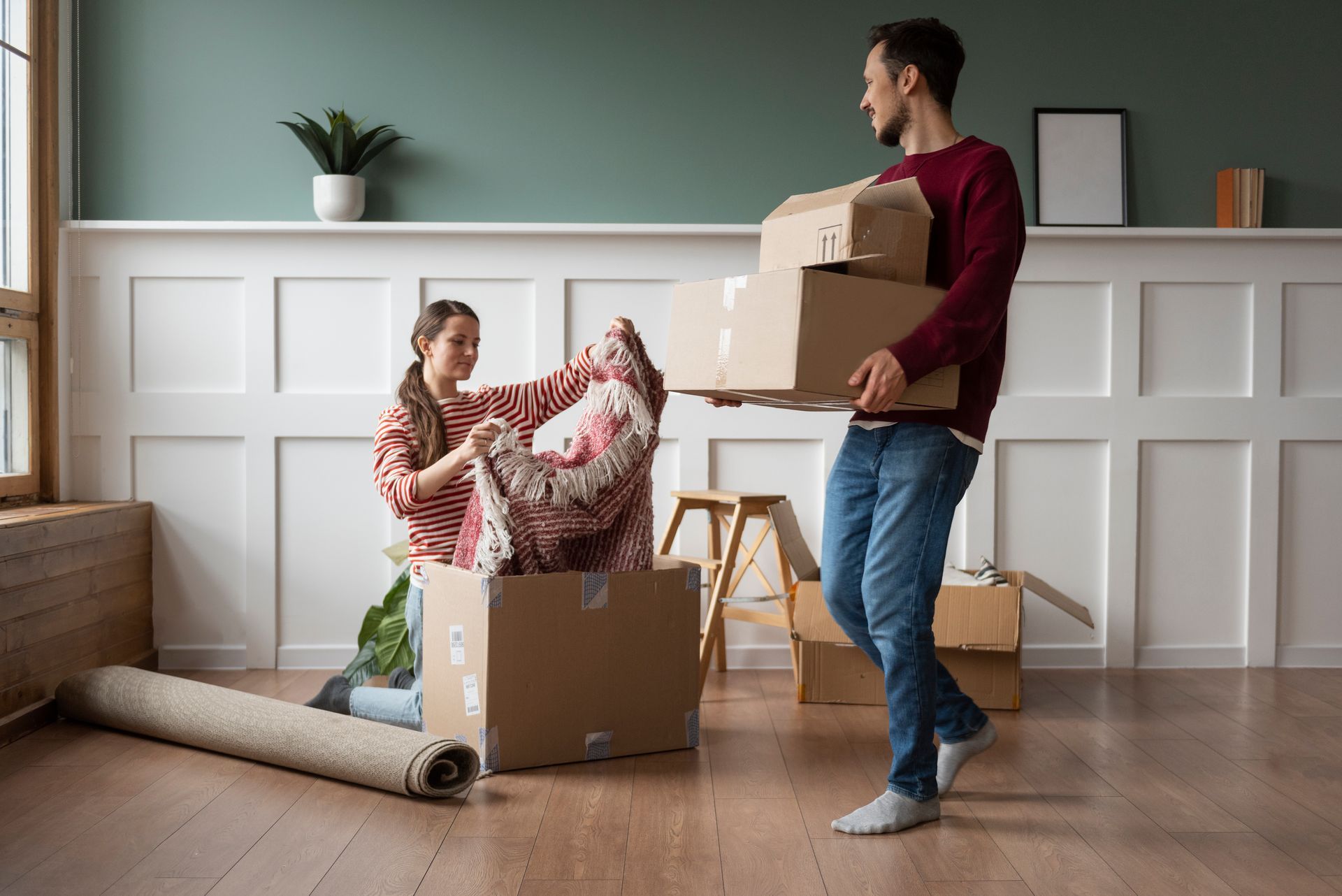 Couple unpacking moving boxes in new home. Man carrying boxes, woman removing blanket.