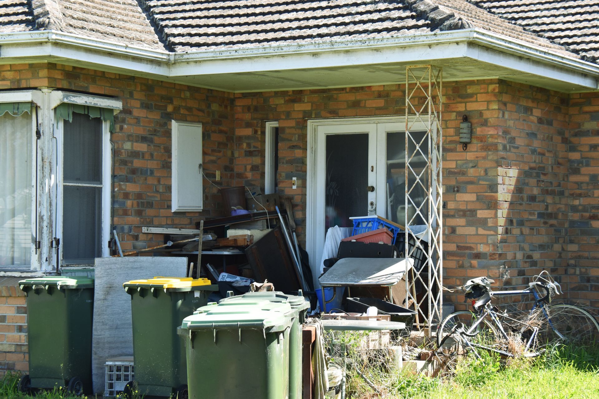 Exterior of a brick house with clutter piled in front of the door, green trash bins, and a bicycle.