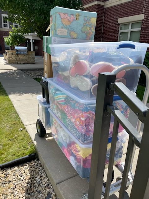 Cart loaded with clear plastic bins and a small trunk, parked on a sidewalk next to a building.