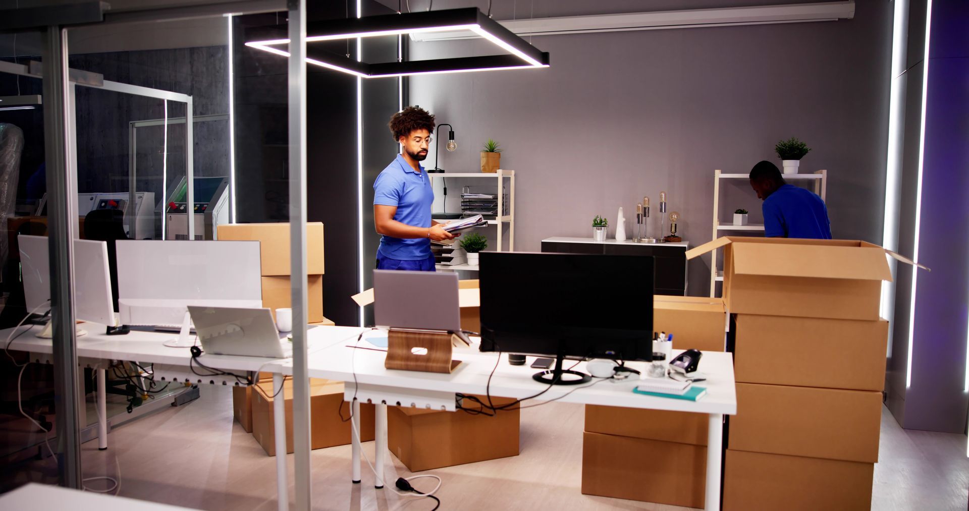 Two people packing cardboard boxes in a modern office space, lit by overhead lights. Desks and monitors are visible.