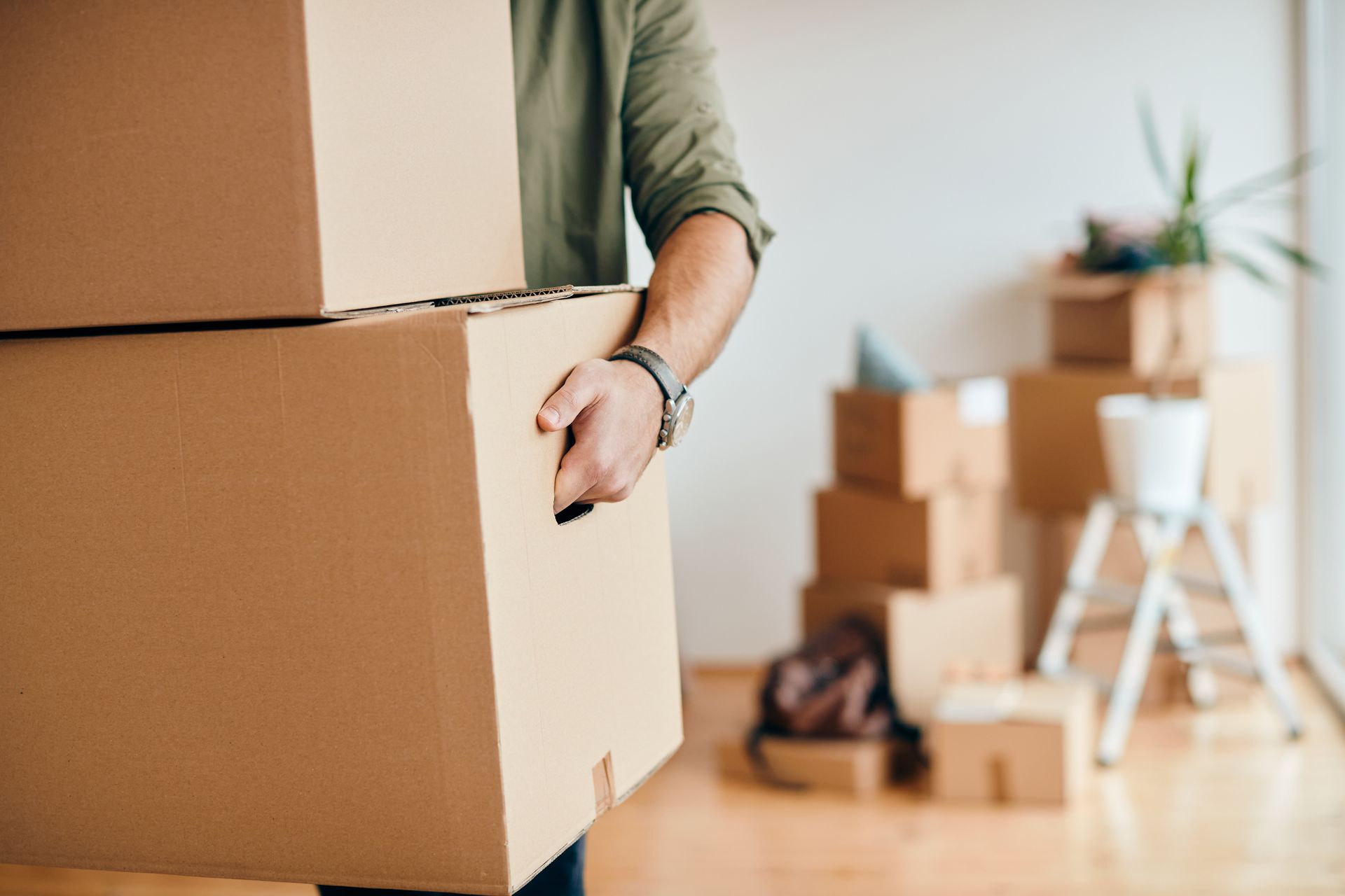 Person carrying two cardboard moving boxes. Boxes, some stacked, fill the room.