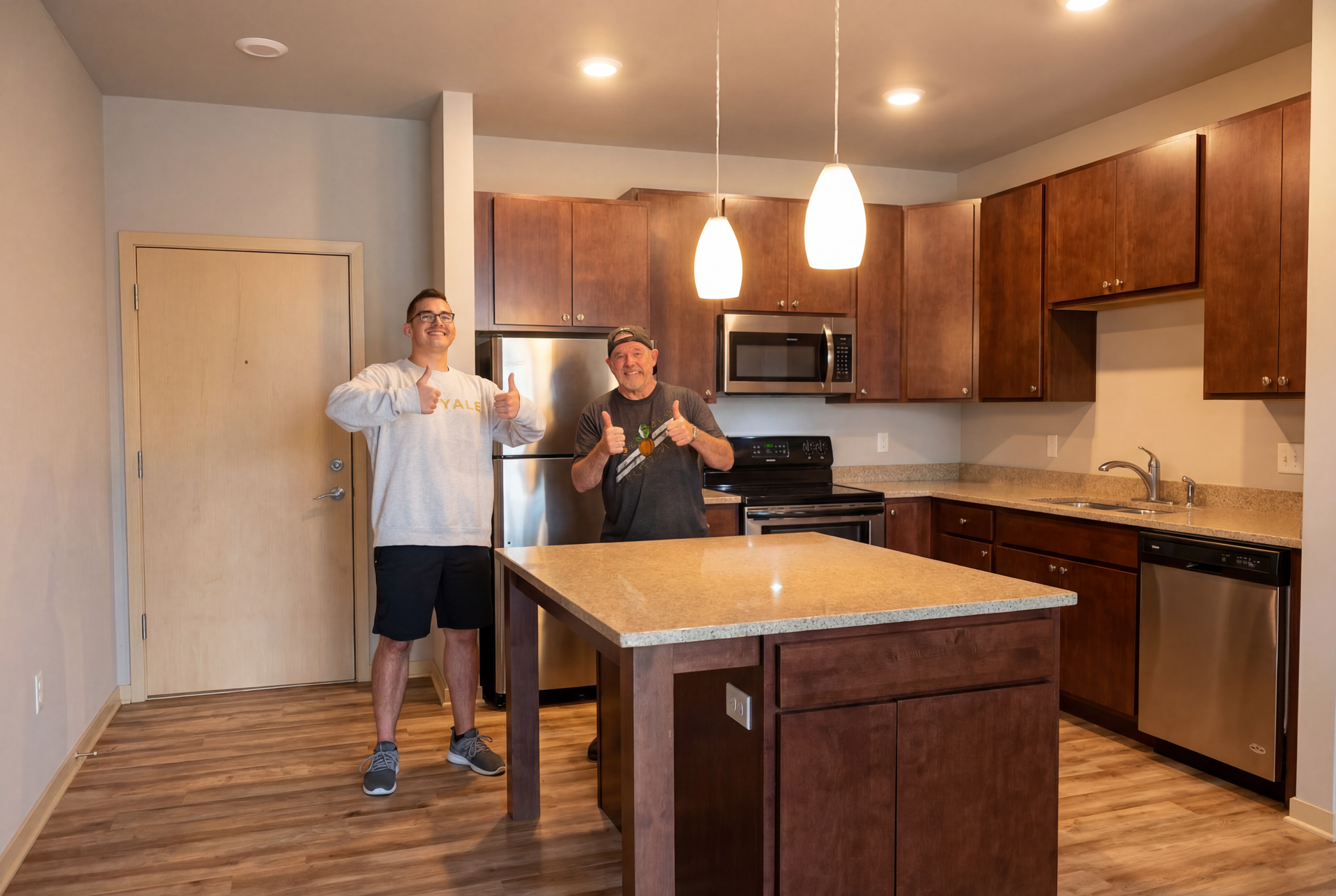 Two men in a new kitchen, giving thumbs up. Brown cabinets, island with granite countertop, stainless steel appliances.