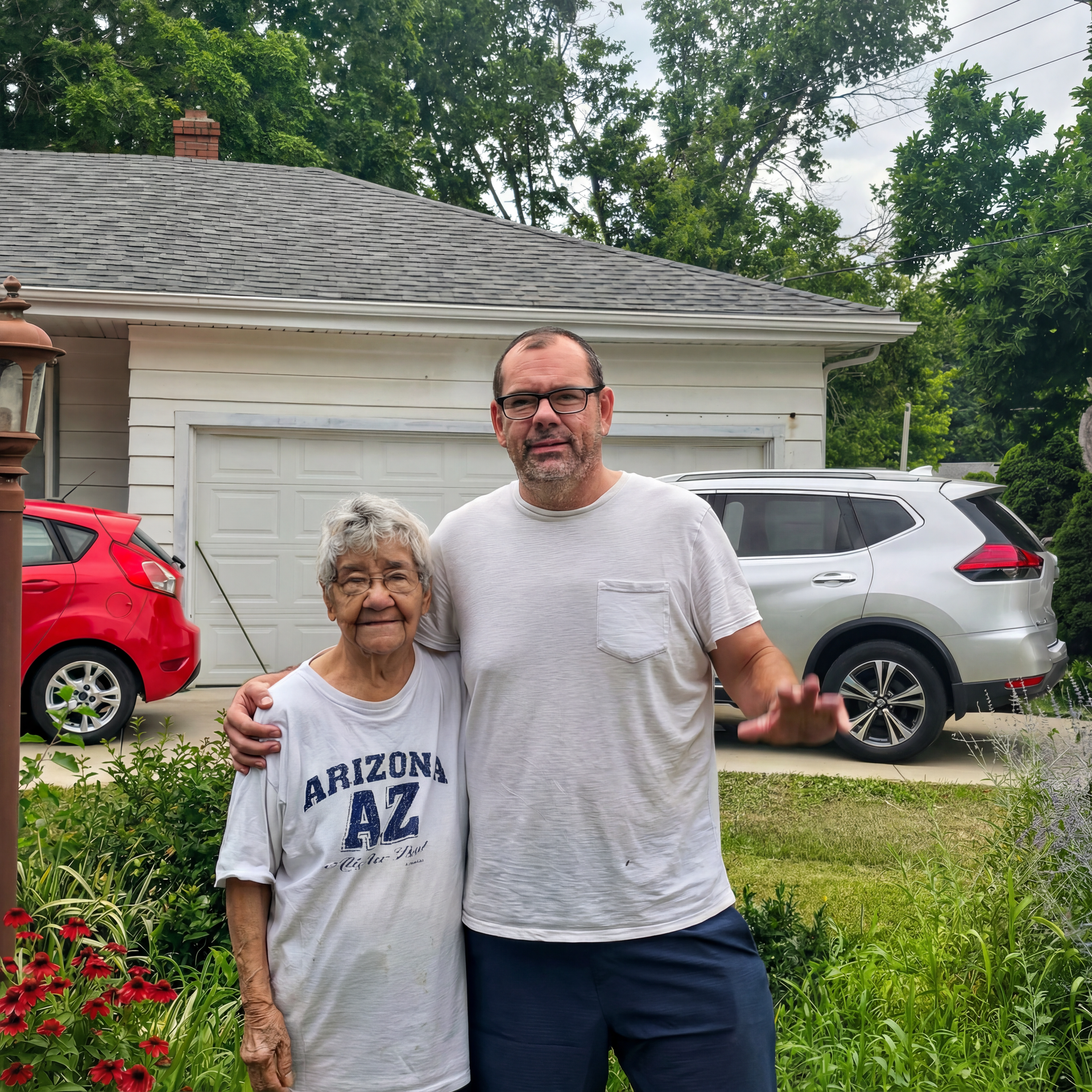 Man with arm around an older woman, standing in front of a house, car in background.