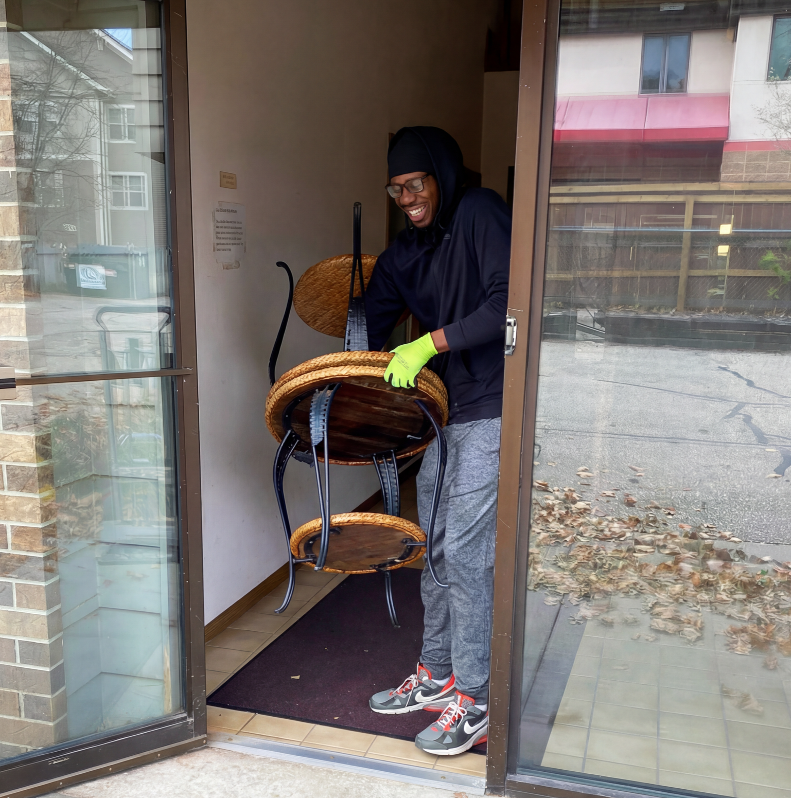 Person carrying a round, wicker-seat chair through a doorway; wearing gloves, hoodie, smiling.