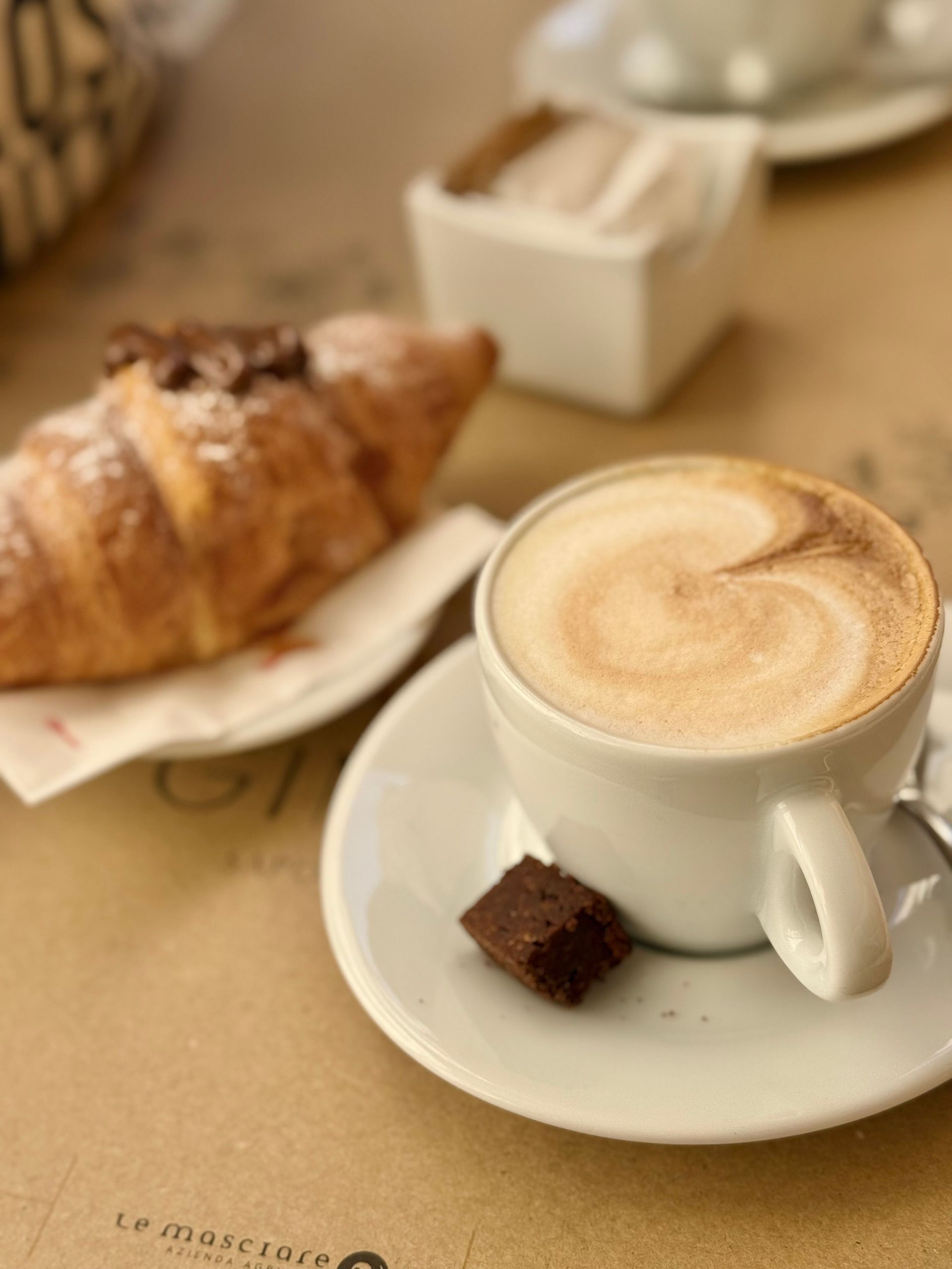 Un cappuccino su un piattino con un croissant su un tavolino da caffè, in una luce calda e soffusa.