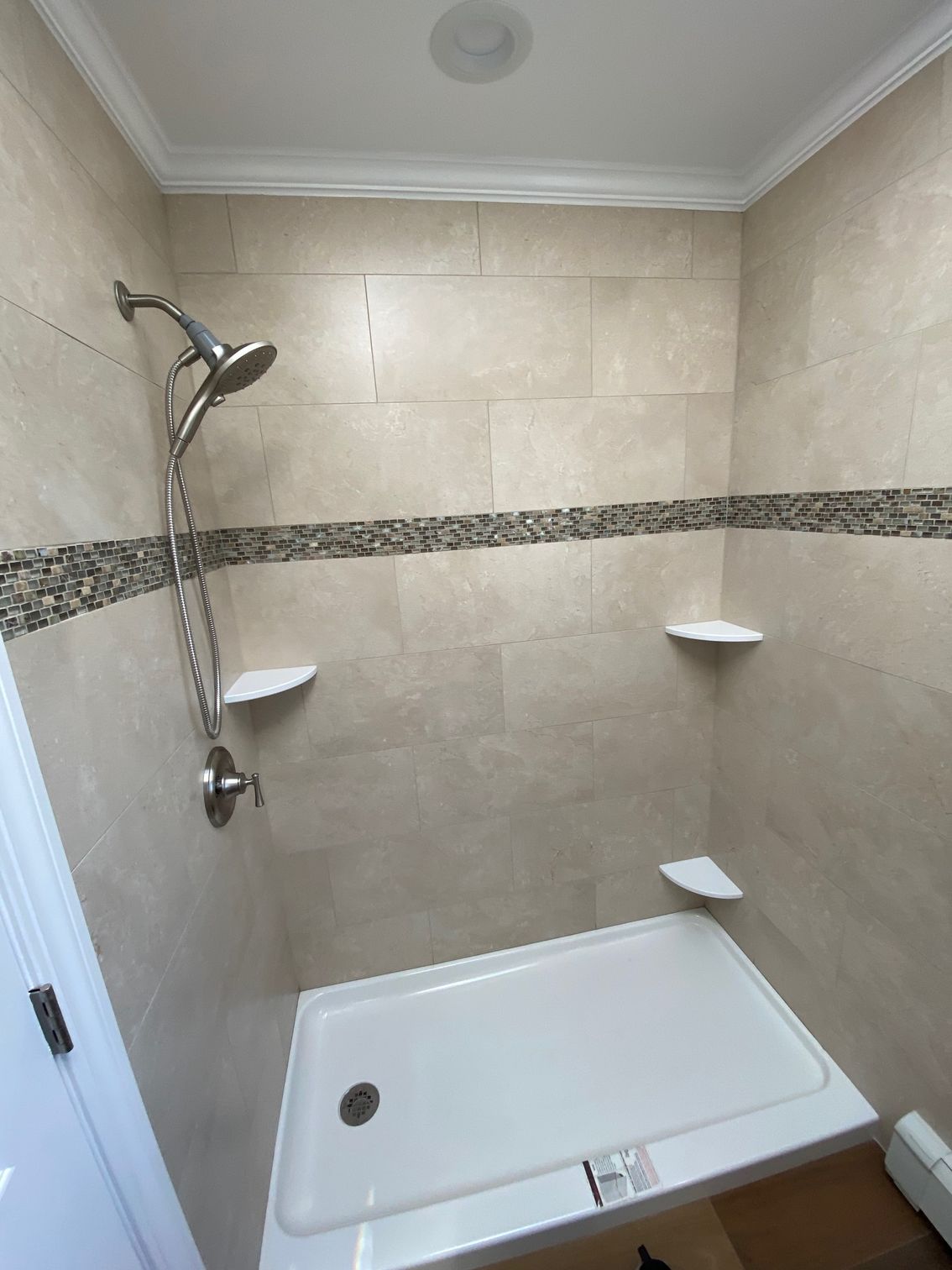 Tiled shower stall with a white base, shower head, and built-in shelves. Beige walls with a decorative stone band.