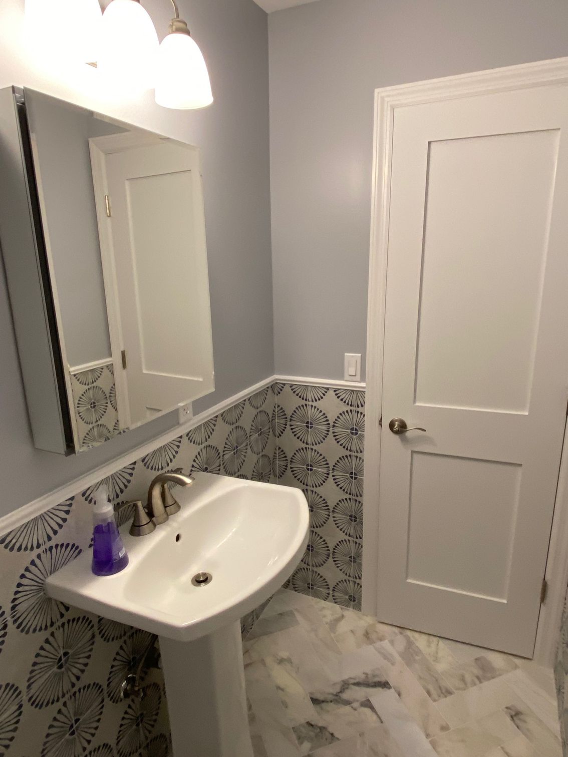 Bathroom with pedestal sink, patterned tile, and a white door. Gray walls, overhead light, and mirror.