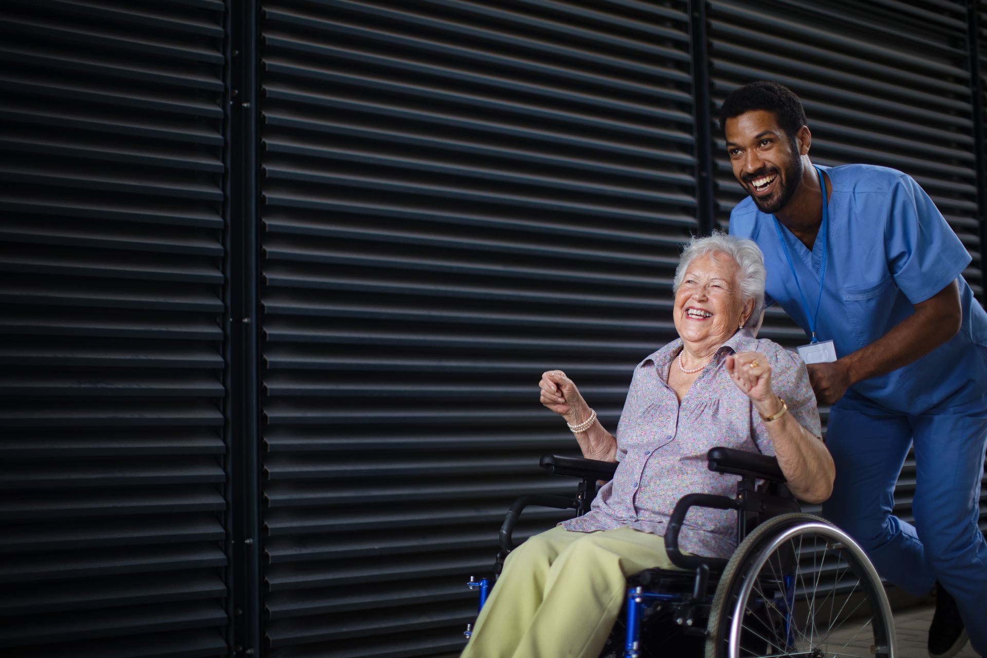 An elderly woman in a wheelchair is laughing with a nurse.