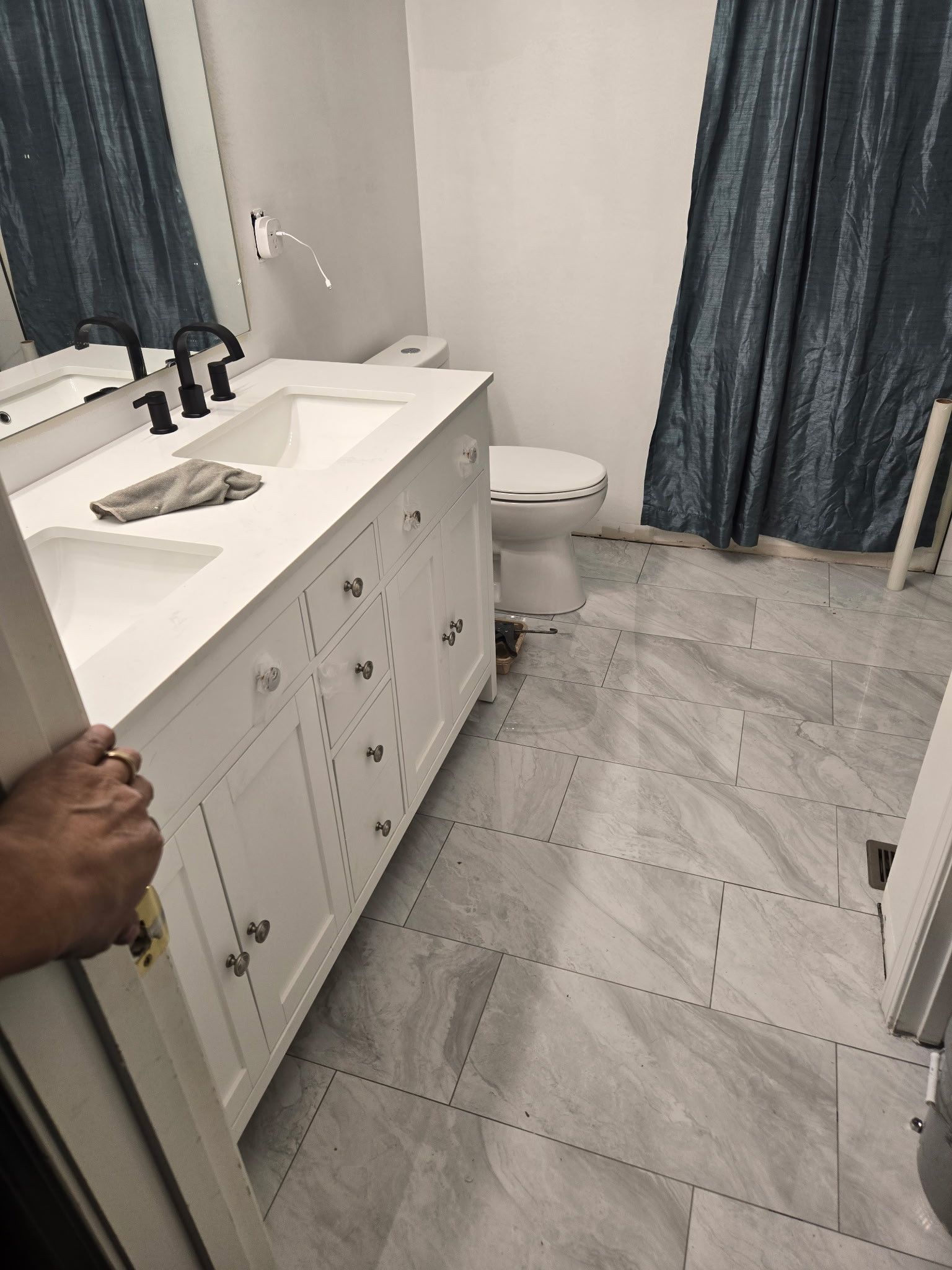 Bathroom with white vanity, toilet, and blue curtains. Door partially open, showing hand.