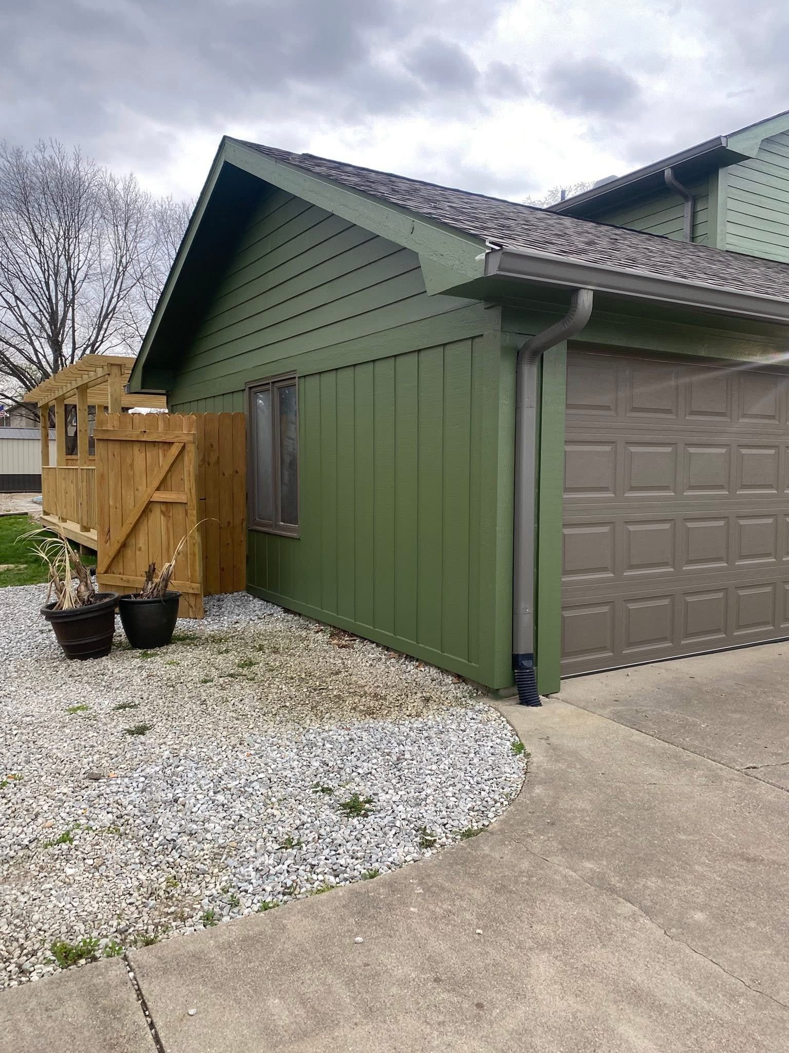 Green-sided building with brown garage door, wooden fence, and concrete walkway, against a cloudy sky.