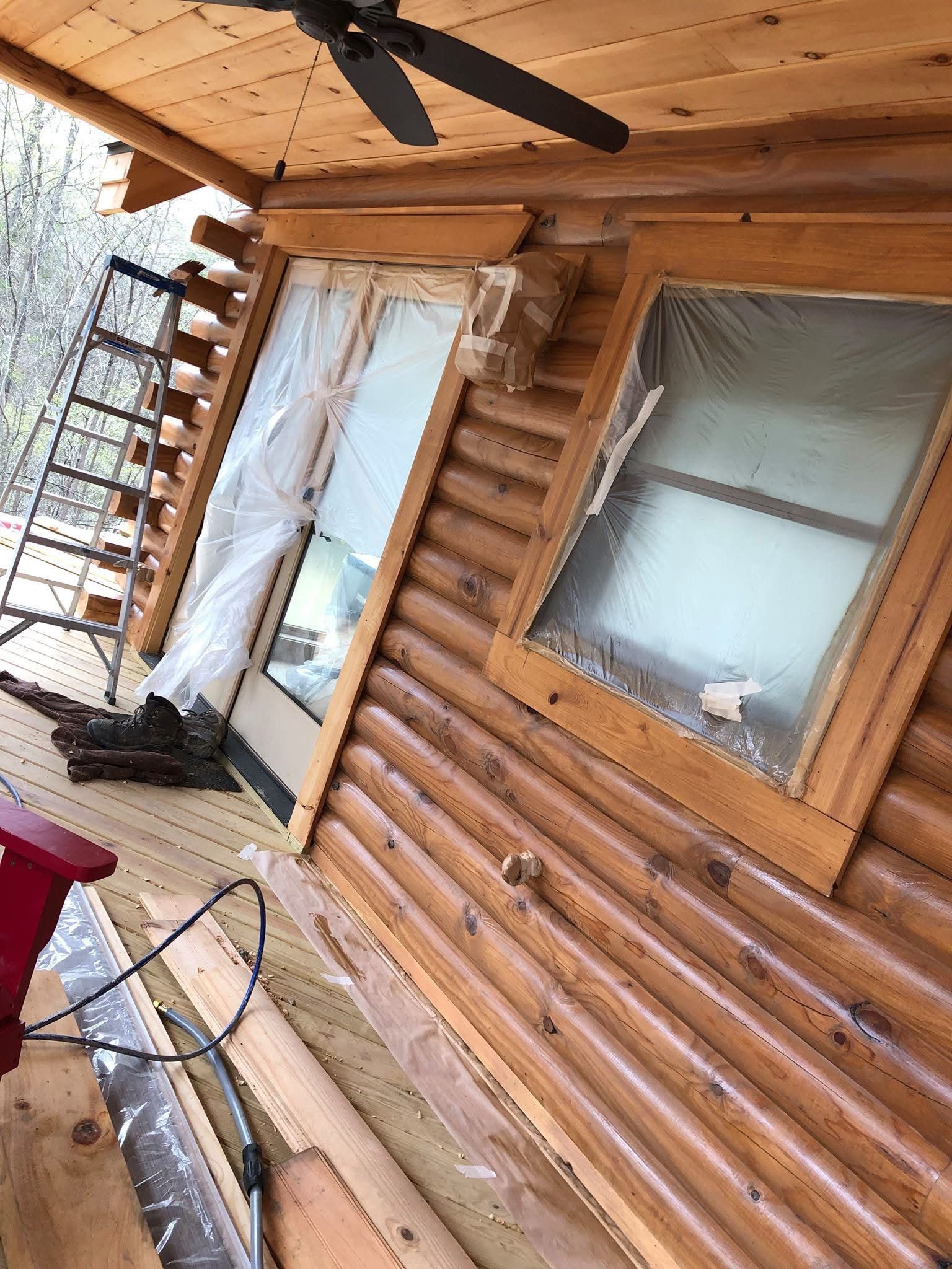 Log cabin exterior with wooden logs. A door and window are covered in plastic. A ladder is present.