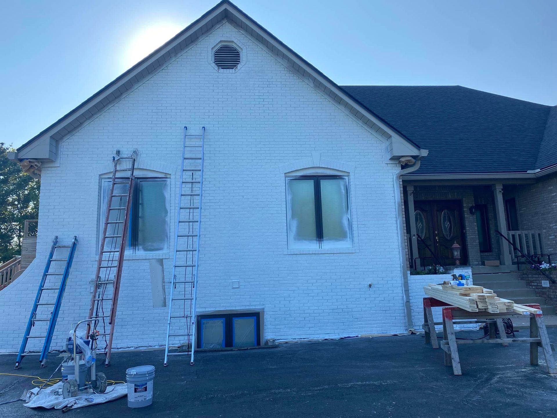 House exterior partially painted white, ladders leaning against the brick. Windows taped for painting.
