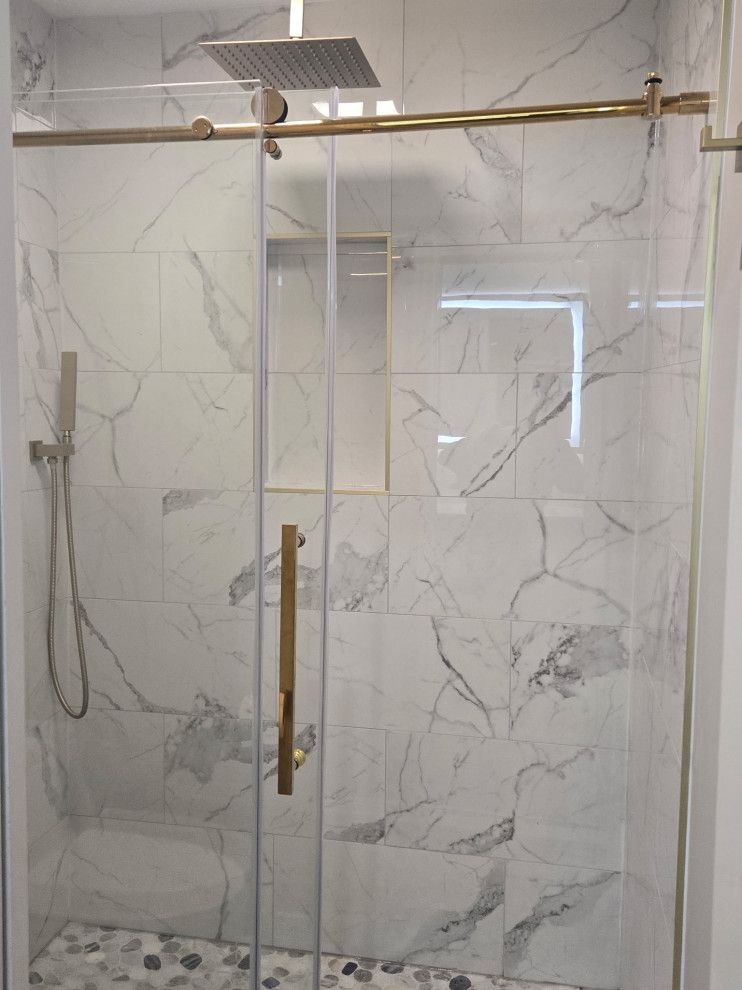 Gold-framed glass shower with marble-look tile, rainfall showerhead, handheld shower, and pebble floor.
