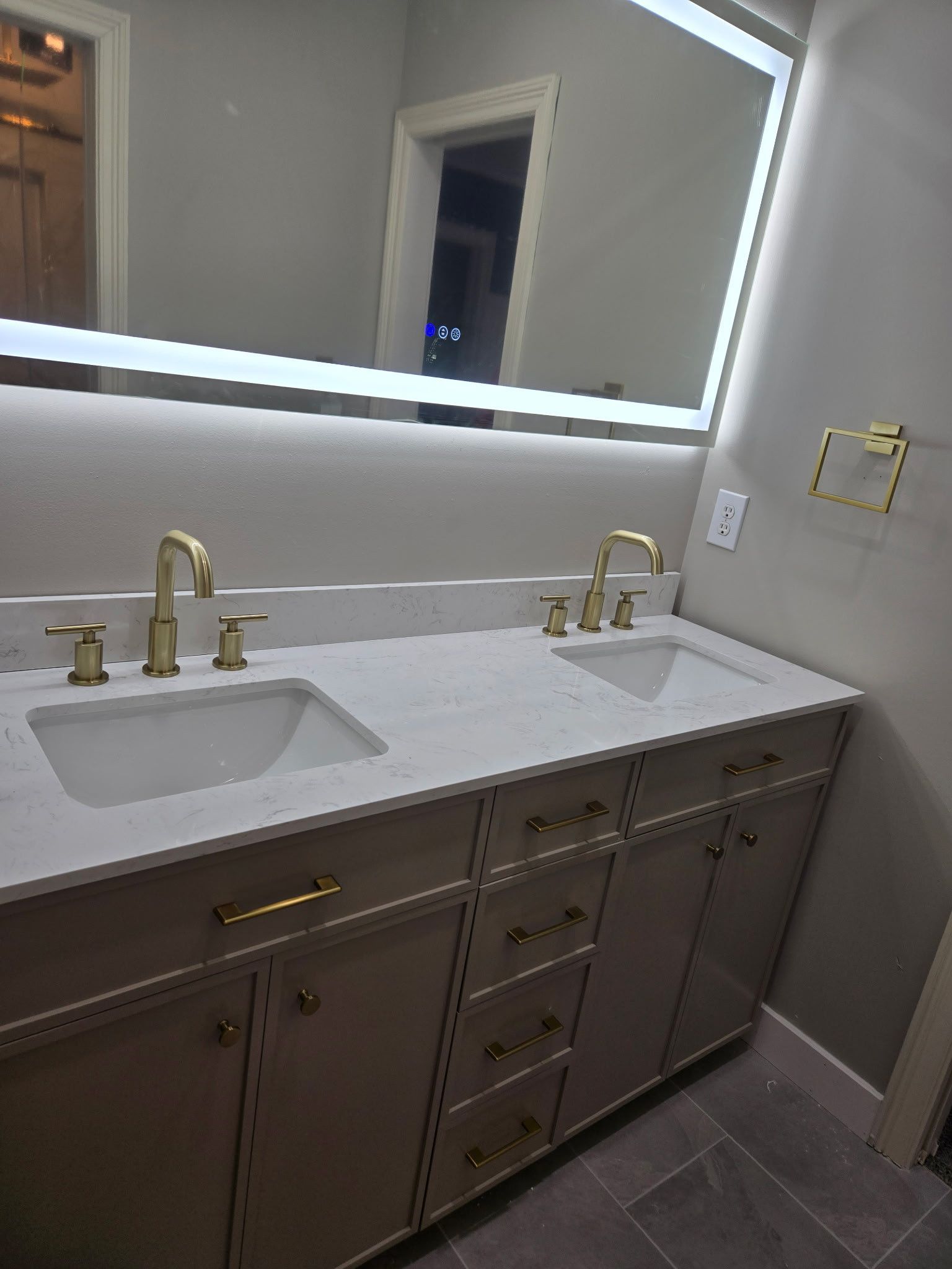 Bathroom with double sinks, gold fixtures, and a large mirror with a lighted frame. Beige vanity.