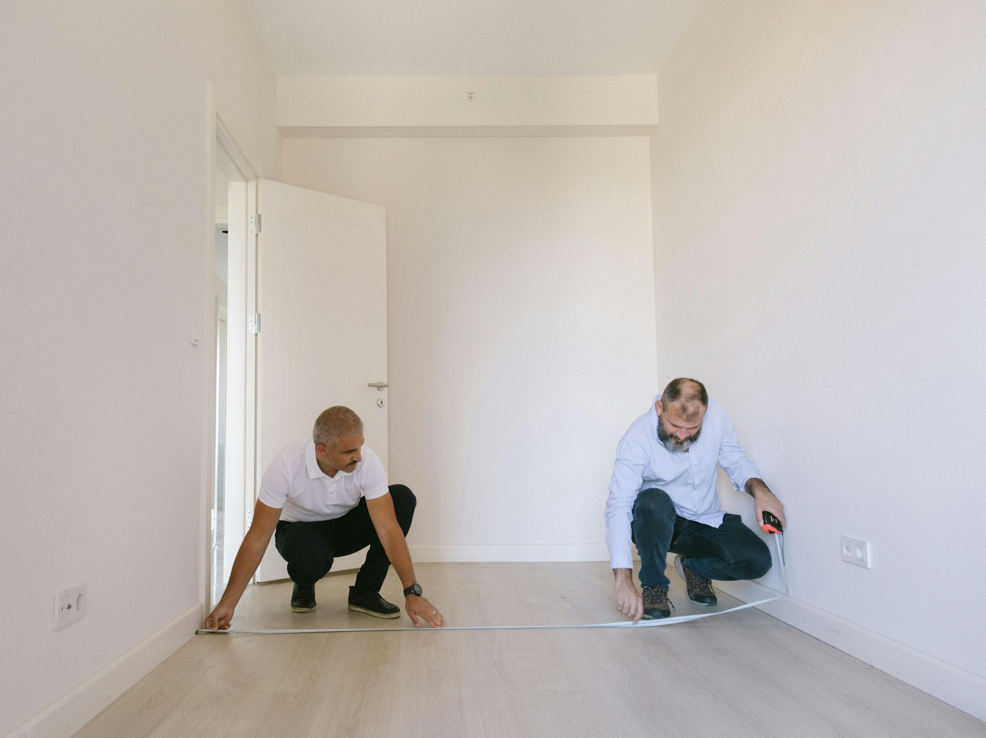 Two people measure a room's floor with a measuring tape, near a door. The room has white walls and light-colored flooring.