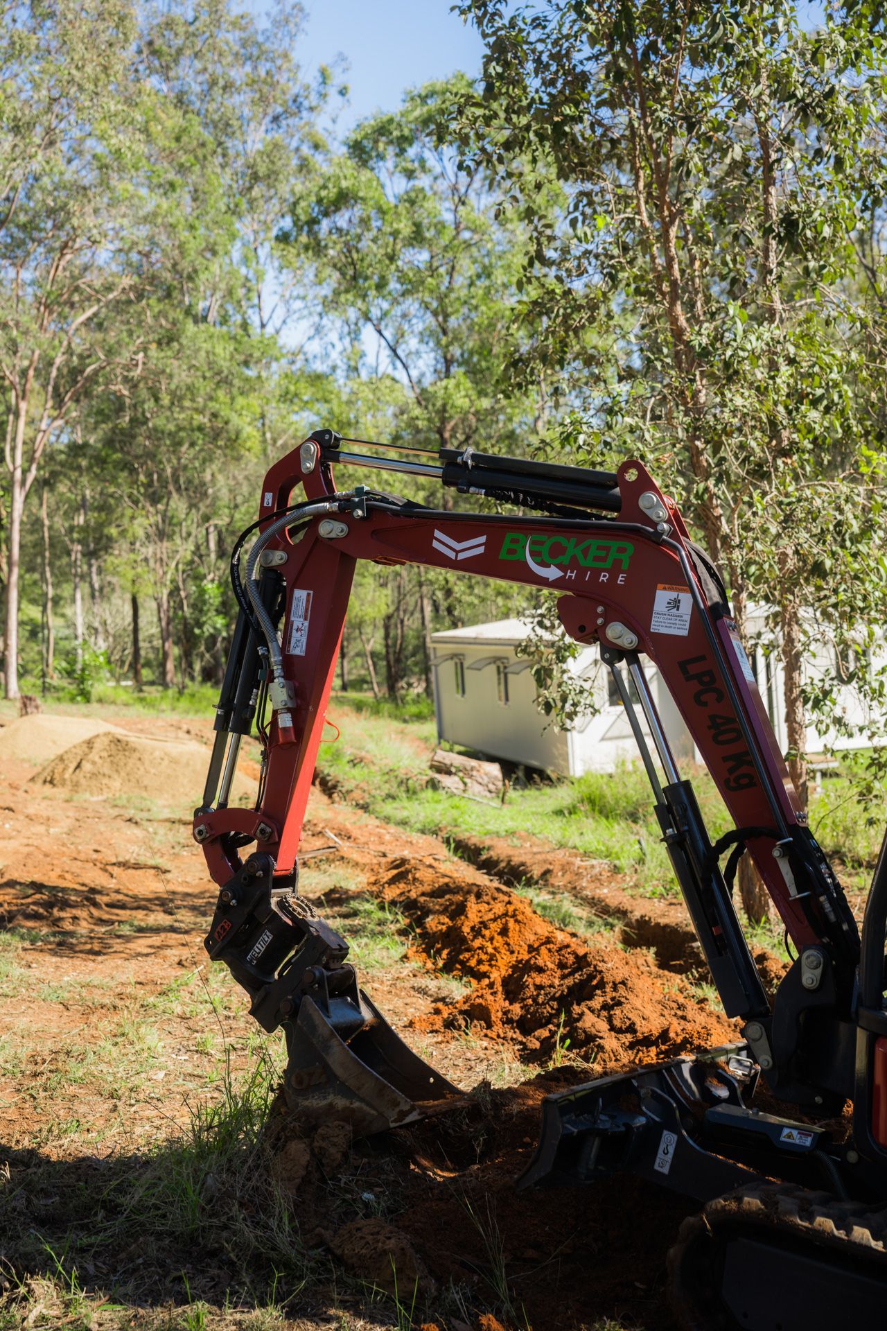 A person operating a small excavator, digging a trench in a wooded area with brown soil.
