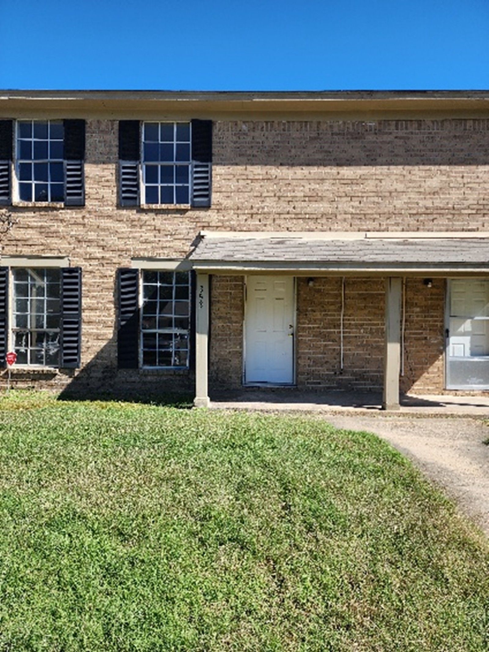 Brick two-story townhome with black shutters, a white door, and a green lawn under a blue sky.