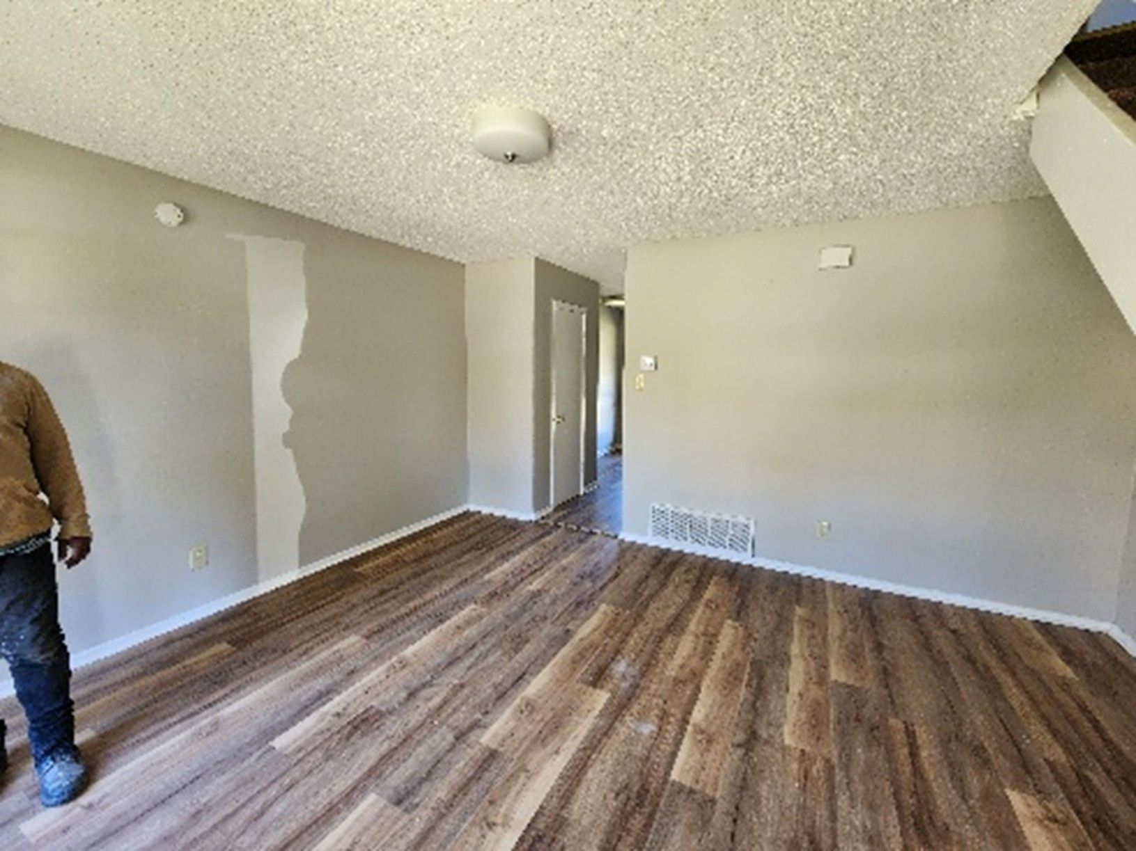 Interior room with wood-look floor, gray walls, doorway, and popcorn ceiling. Man standing in foreground.