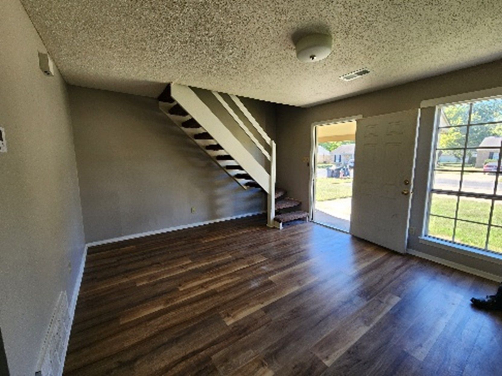 Empty living room with wood floors, stairs, door to the outside, and a window.
