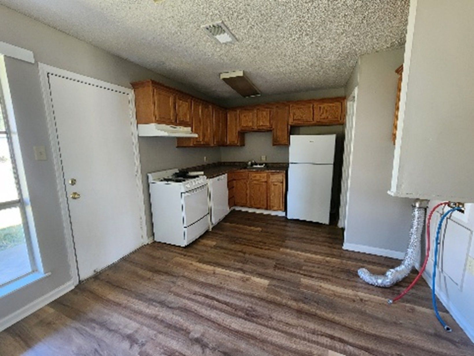 Kitchen with wood cabinets, white appliances, and brown flooring. Door to the left, vent hood over the stove.
