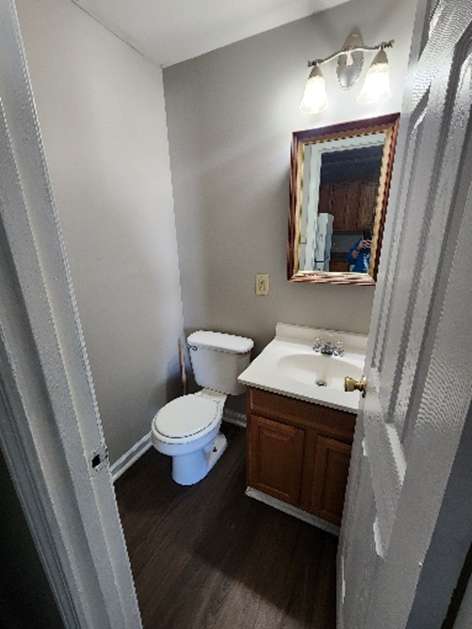 Small bathroom with a toilet, sink, mirror, and wooden cabinet. Grey walls and dark flooring.