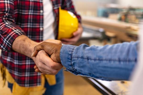 A man in a plaid shirt is shaking hands with a man in a hard hat.