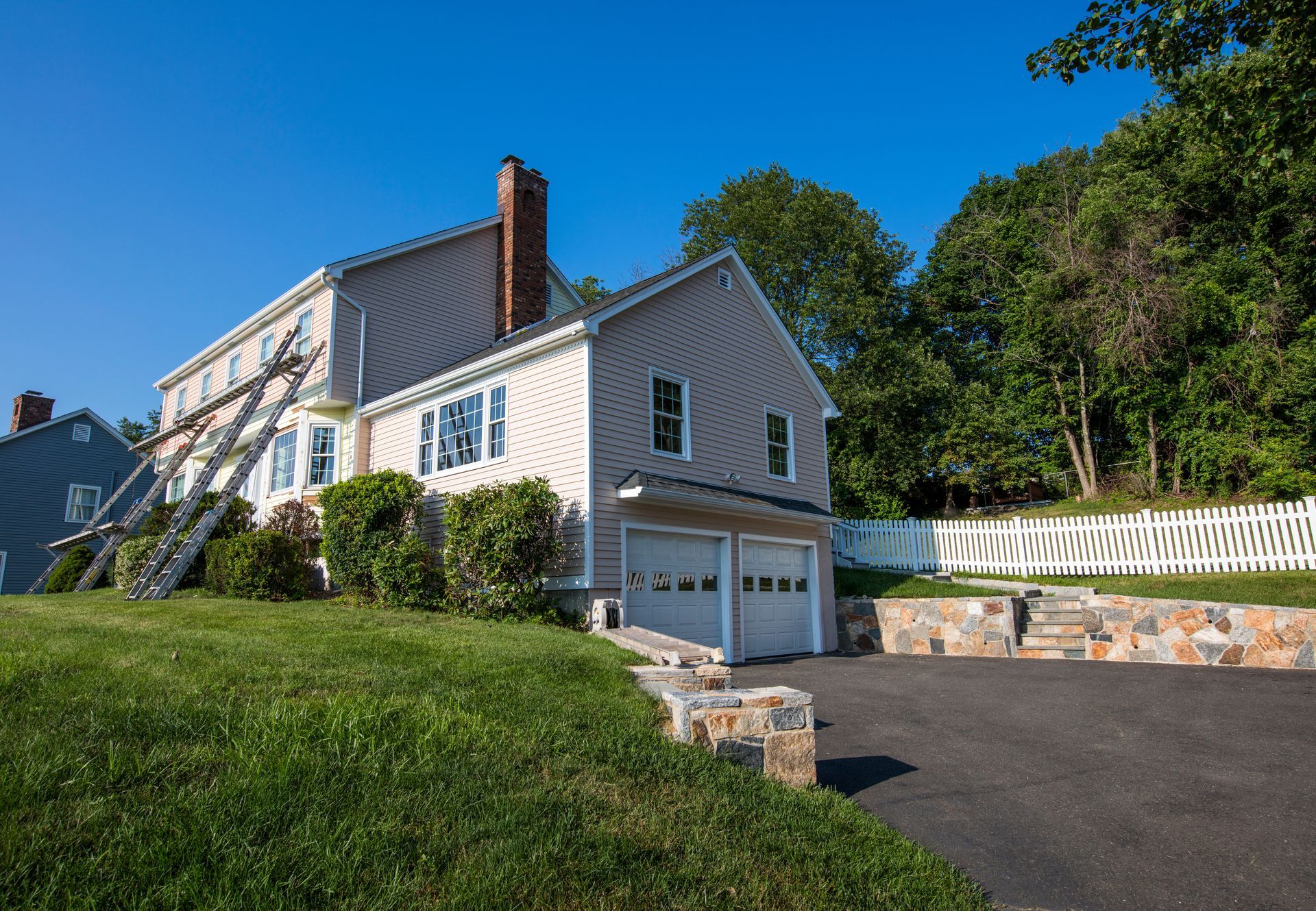 Beige house with a two-car garage, ladder against the side, and a driveway on a sunny day.