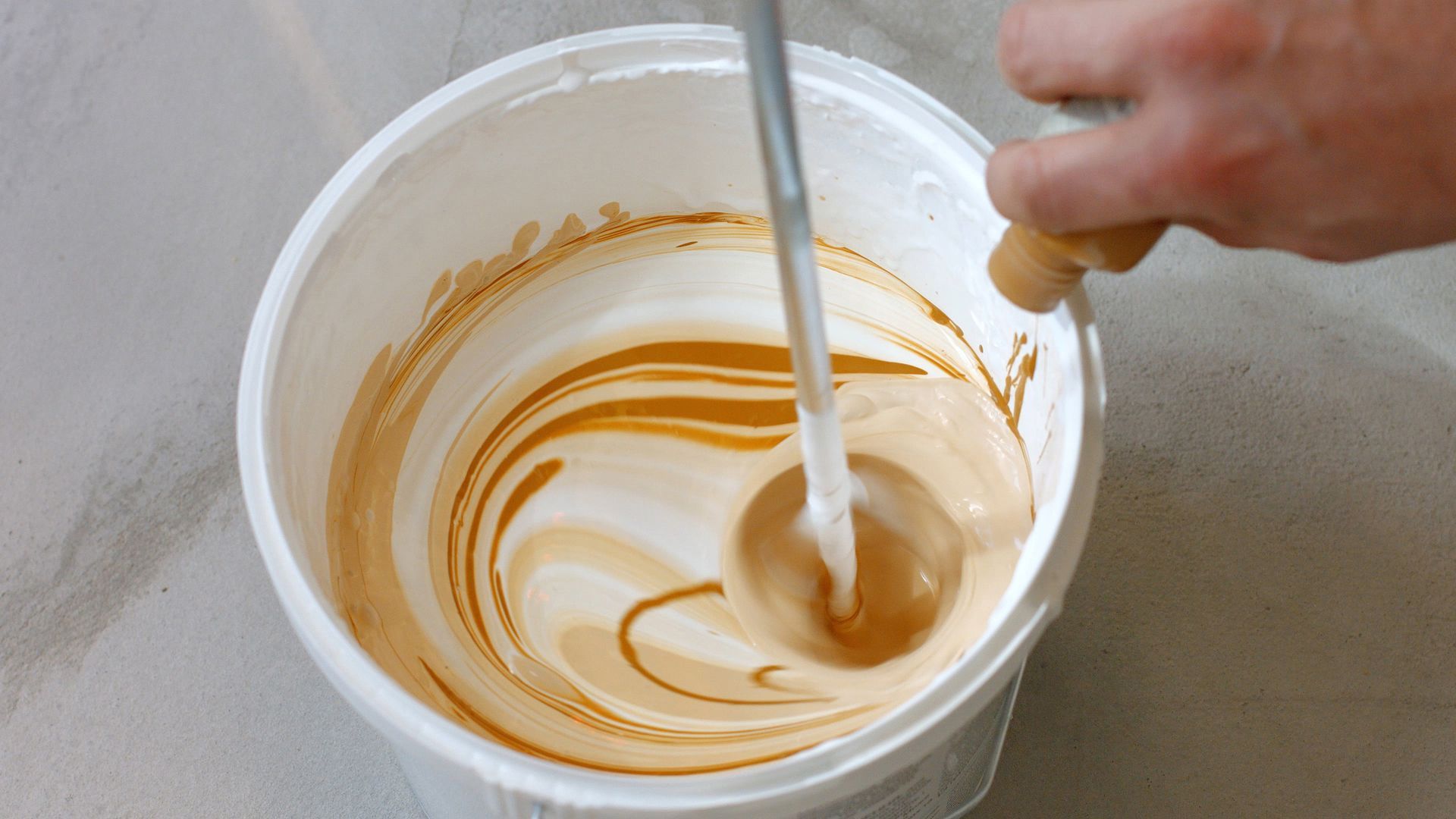 Person mixing tan-colored liquid in a white bucket with a handheld mixing tool.