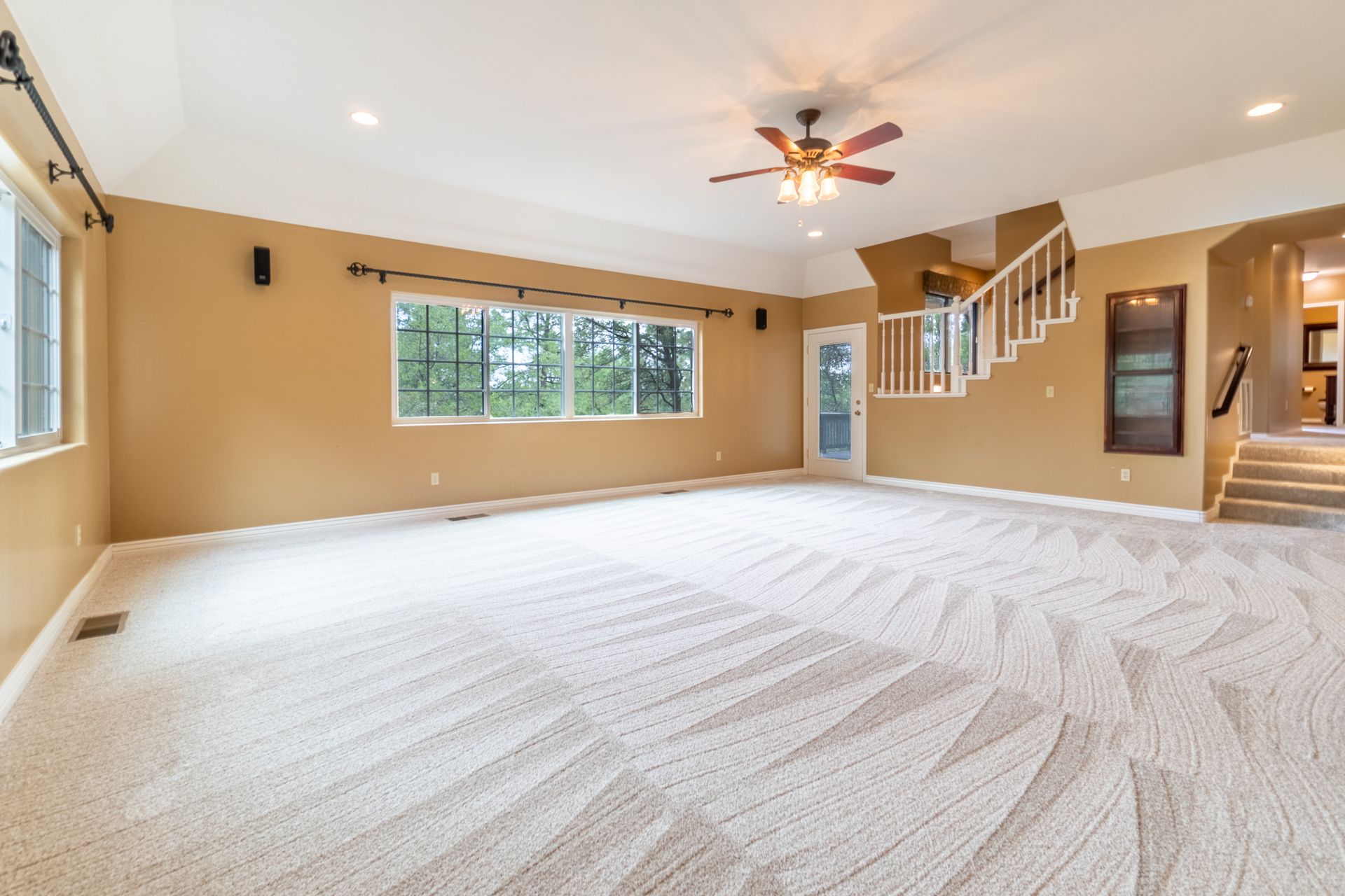 Spacious room with beige walls, patterned carpet, large window, stairs, and a ceiling fan.