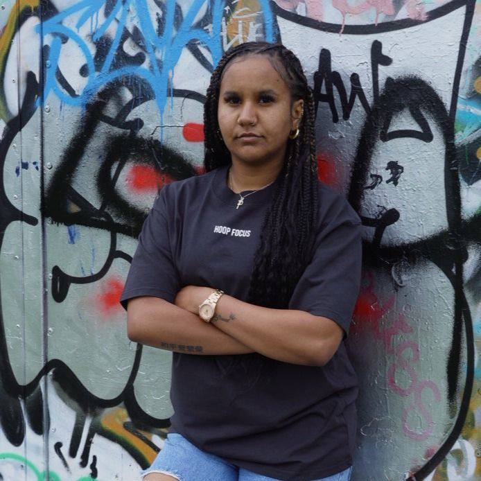 Woman with arms crossed, in front of graffiti. Wearing a dark shirt and blue jeans.