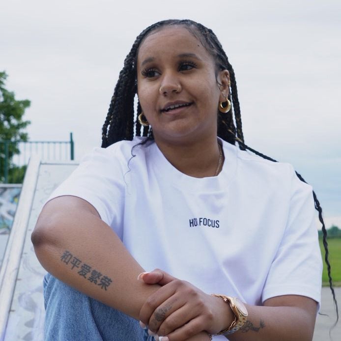 Woman in white shirt with arm tattoo, sitting outside at skate park.